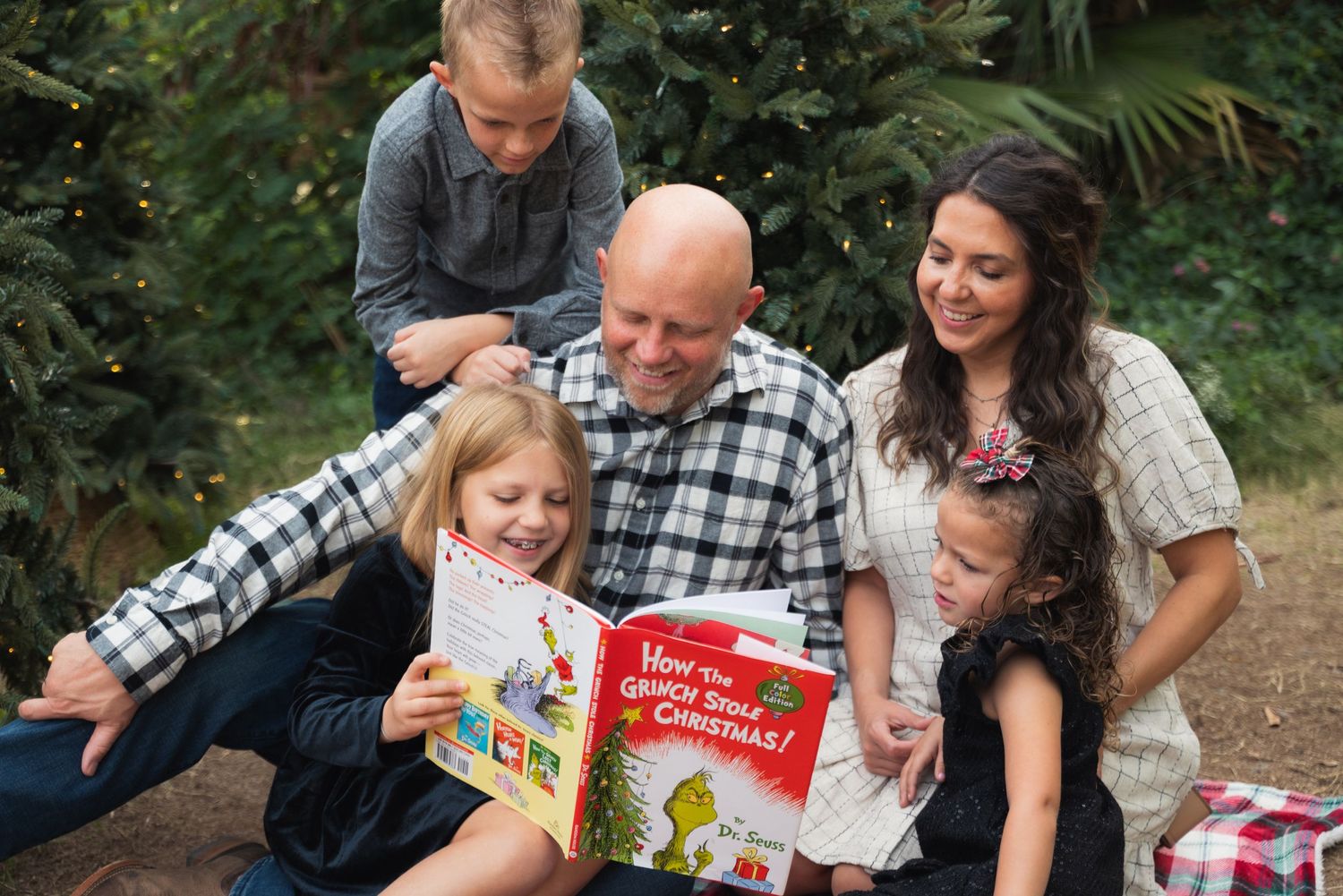 Family of five joyfully reading a Dr. Seuss Christmas book together outdoors near a festive tree.