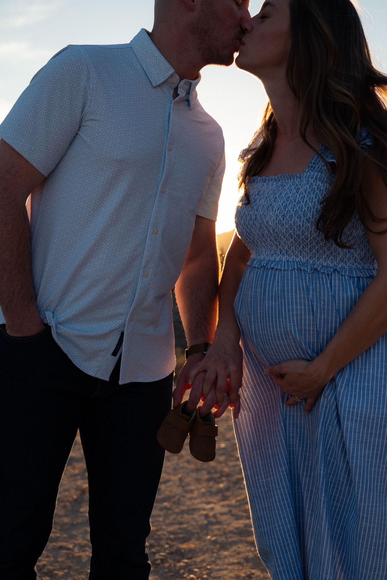 A couple holds hands during sunset at the beach, with the woman wearing a blue dress and the man in a white shirt.