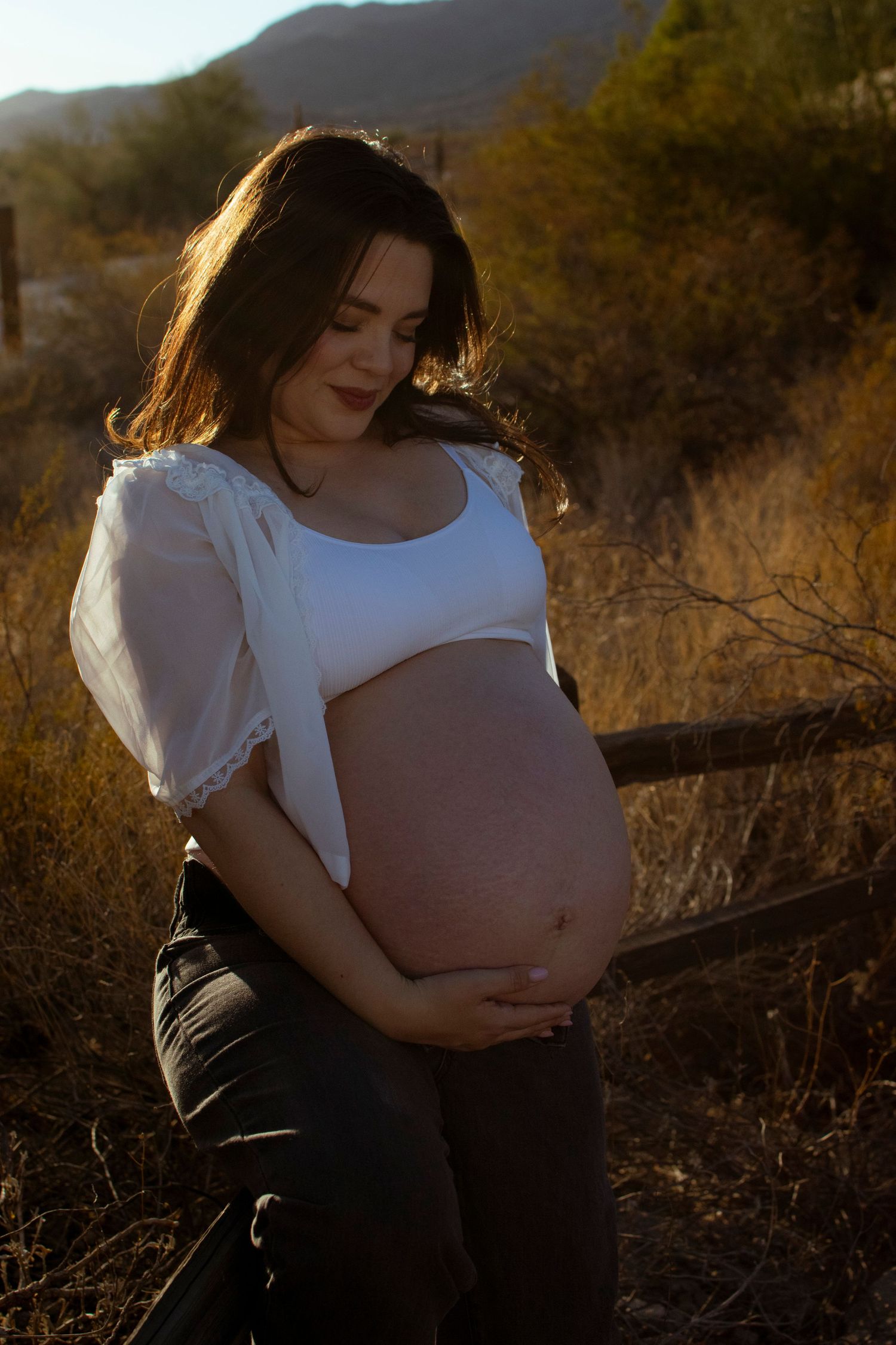An expectant mother in white holds her belly while standing in golden desert light.