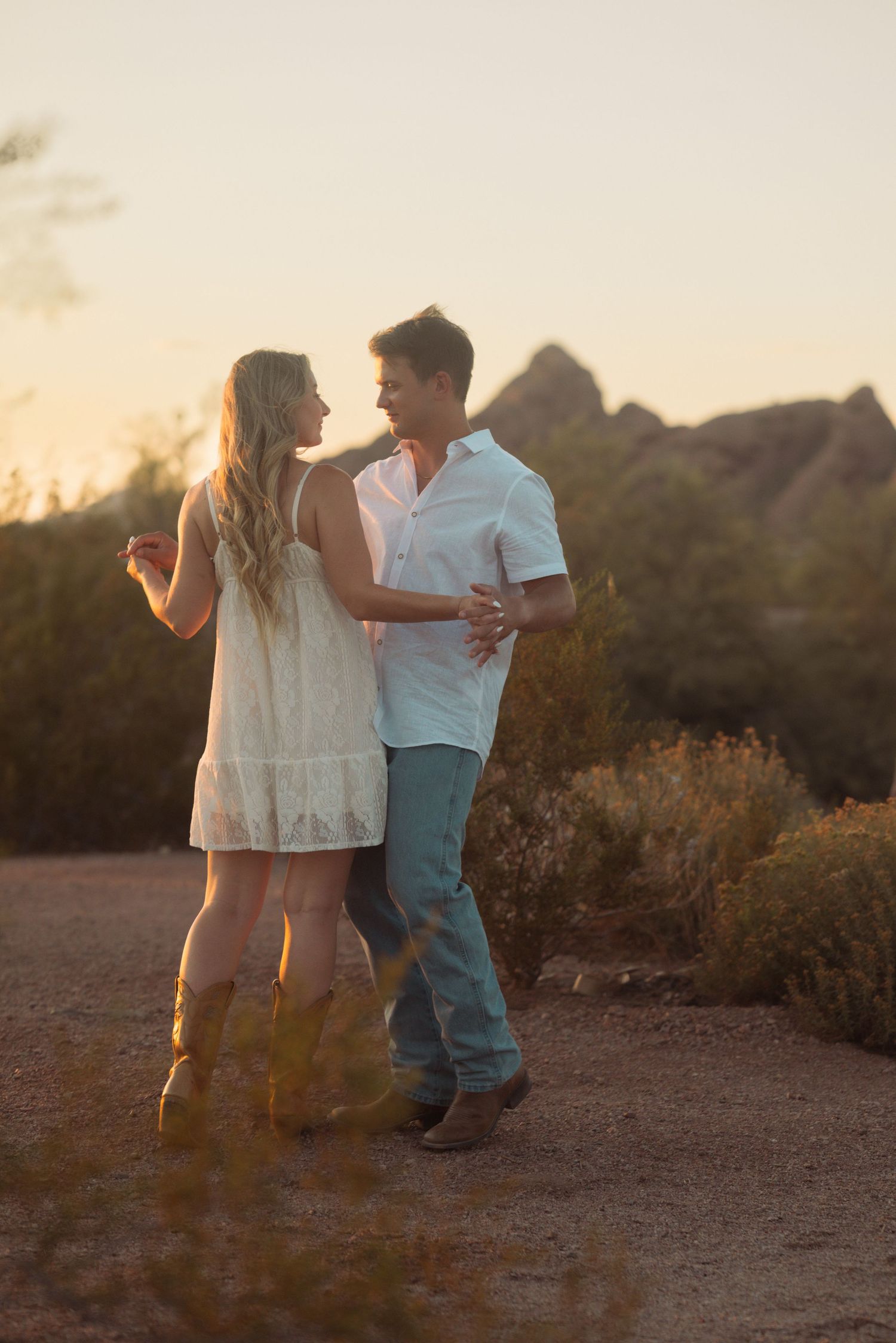 Couple dancing outdoors at sunset in desert landscape, woman in white dress and man in blue jeans embracing romantically.