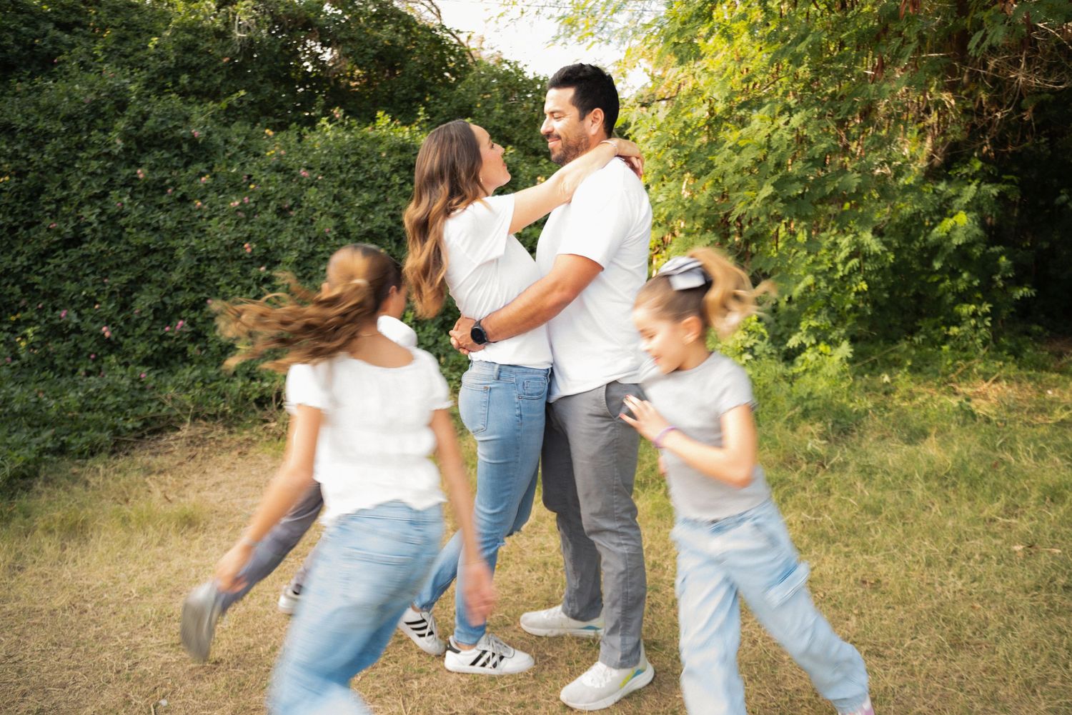 Family in white shirts and jeans embrace outdoors, with children playing around parents near green trees.