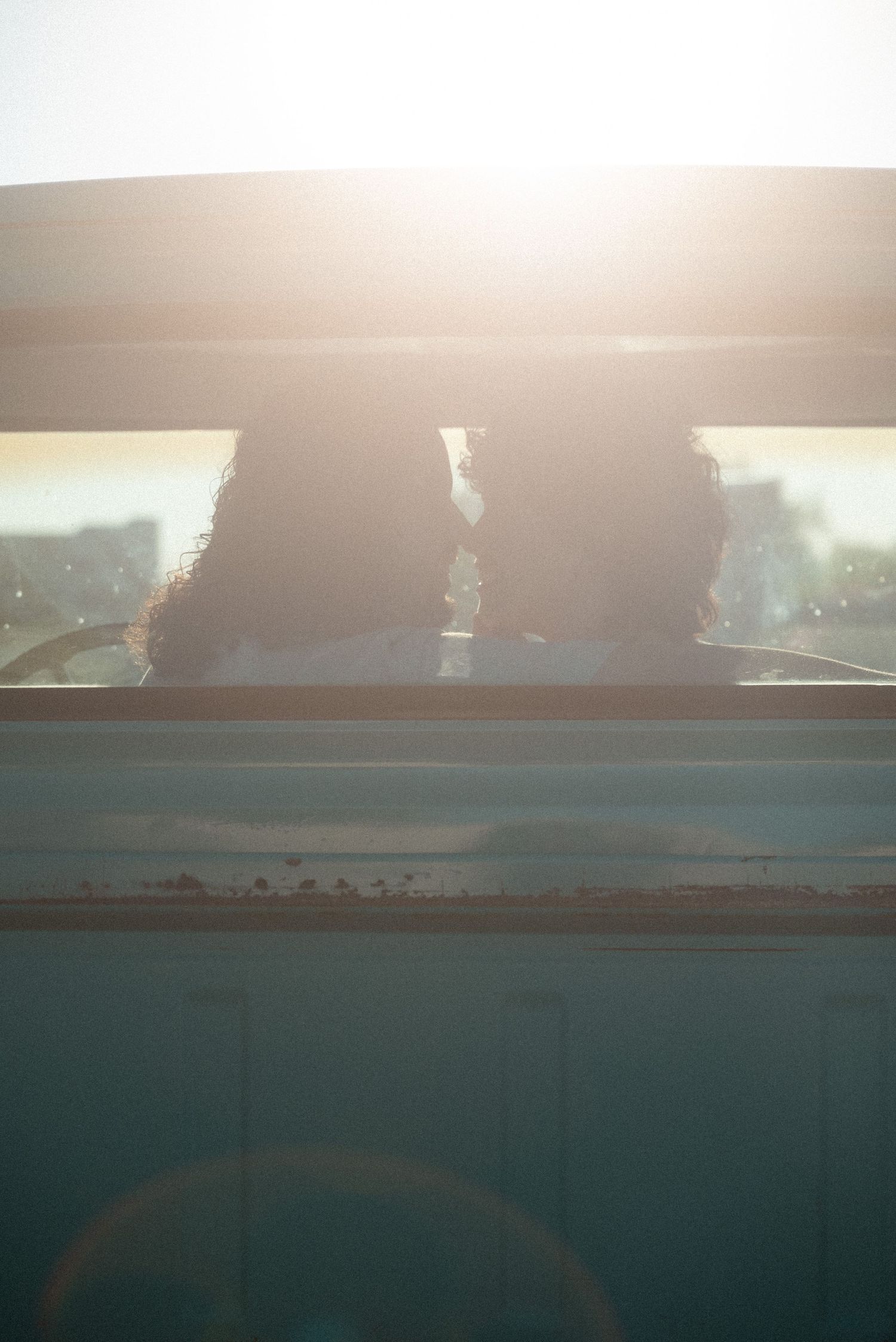 Two people with curly hair sit inside a vintage van, silhouetted against bright golden sunlight streaming through the window.