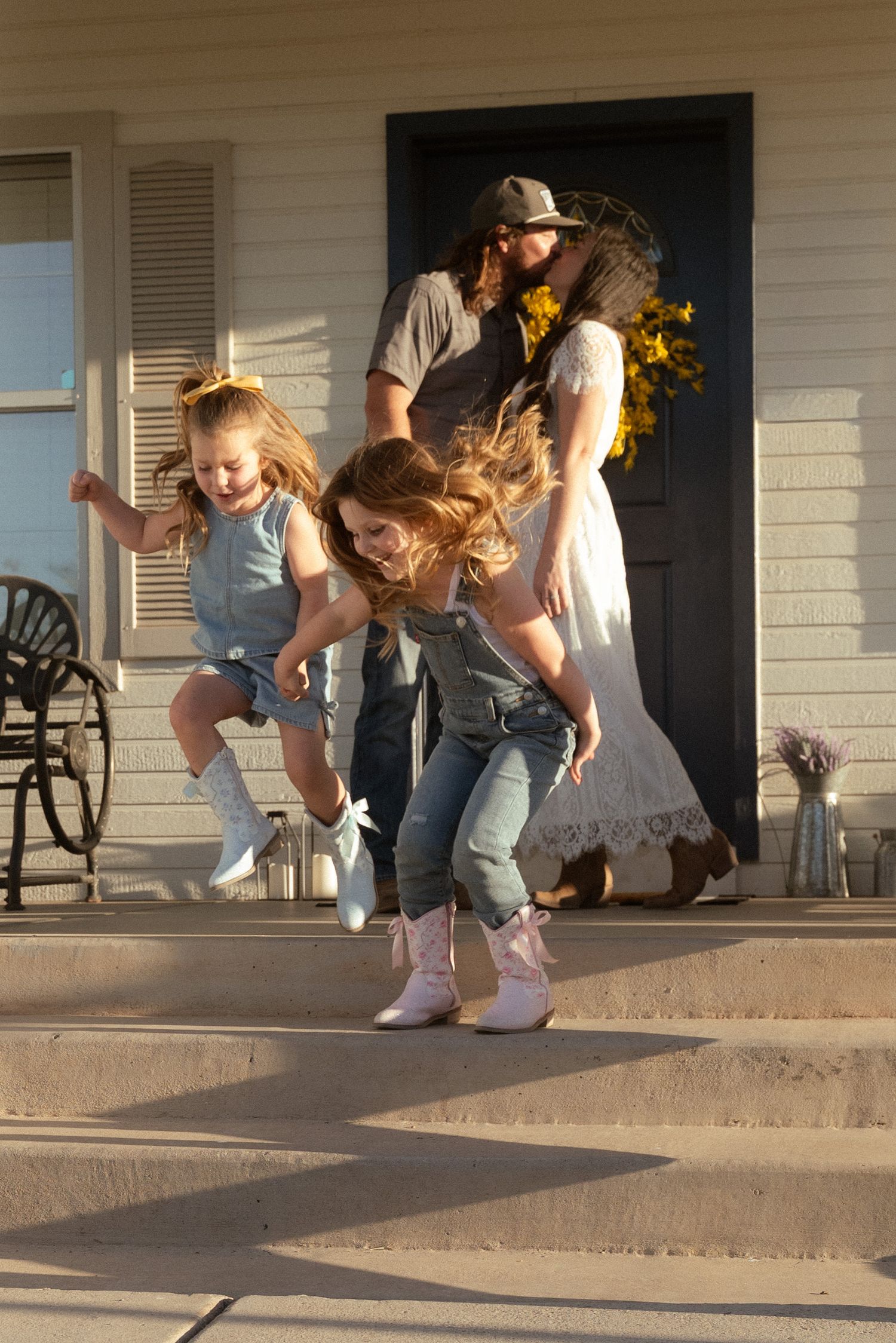 Two young girls in denim and cowboy boots play on porch steps while parents watch from a sunlit farmhouse entrance.