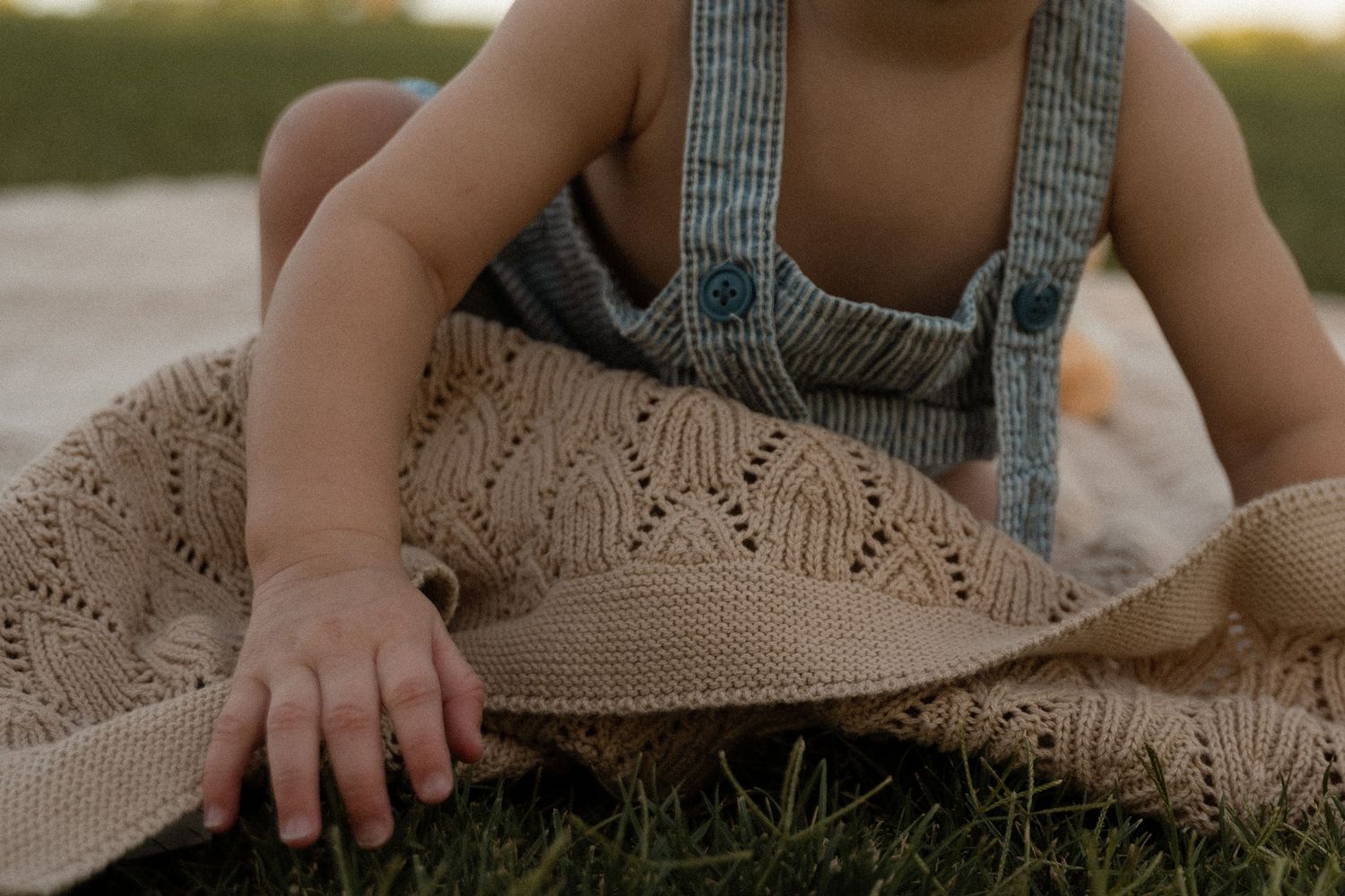 Baby in striped overalls sitting on grass with a knitted beige blanket and straw hat.