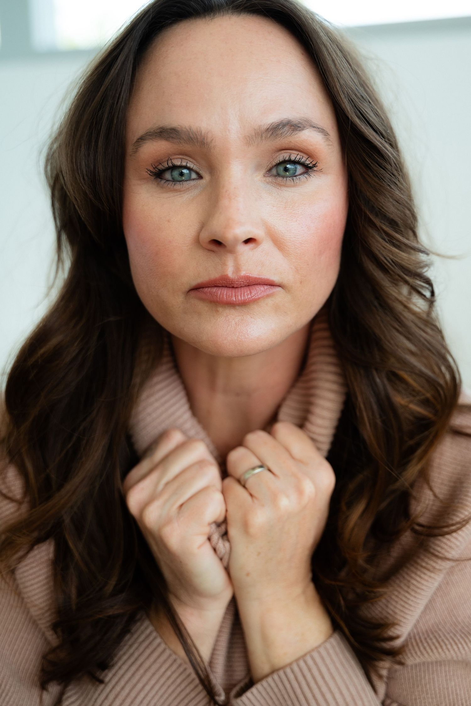 A portrait of someone with wavy brown hair and blue eyes wearing a beige turtleneck sweater.