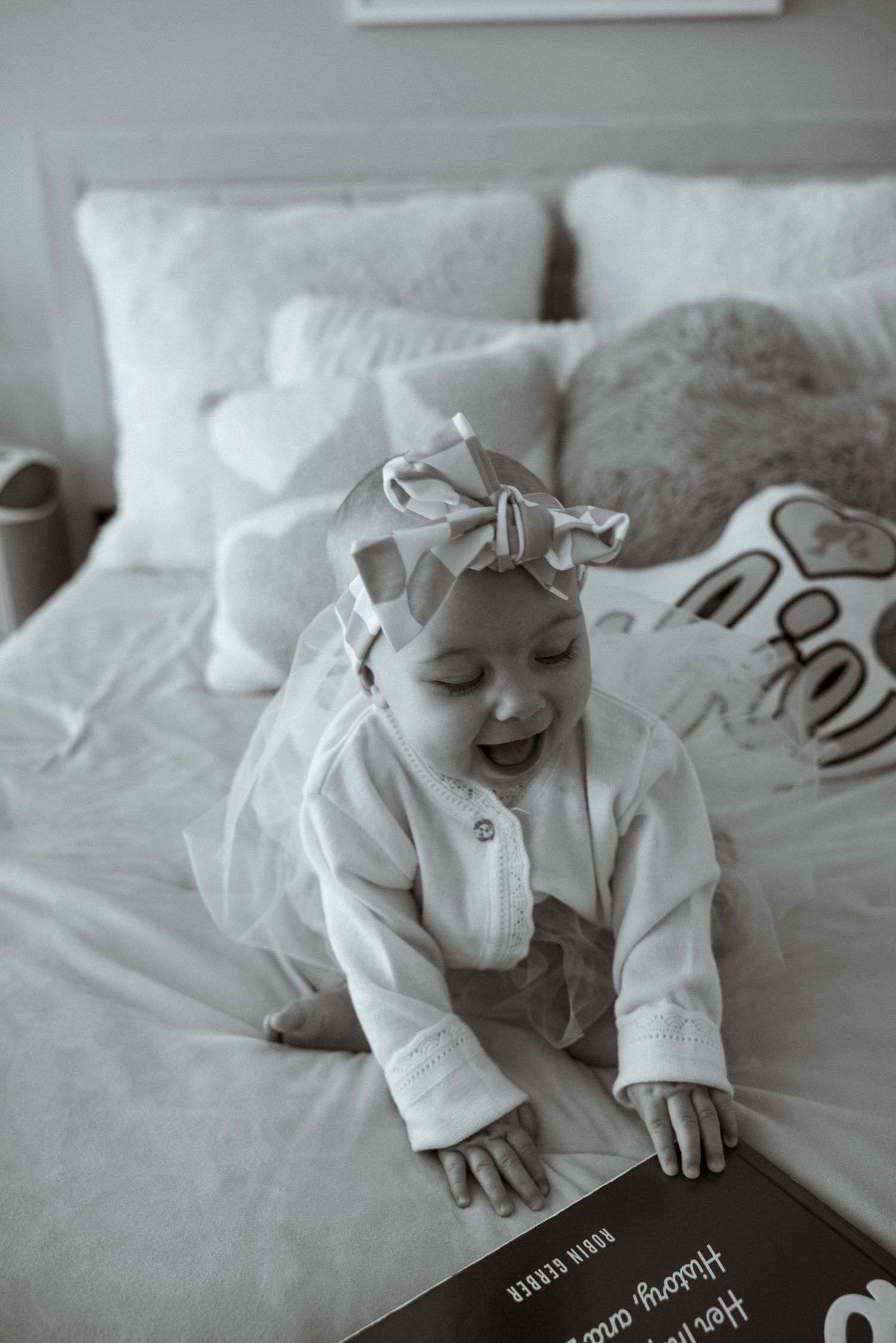A small baby wearing white smiles while lying on a cozy bed with pillows.