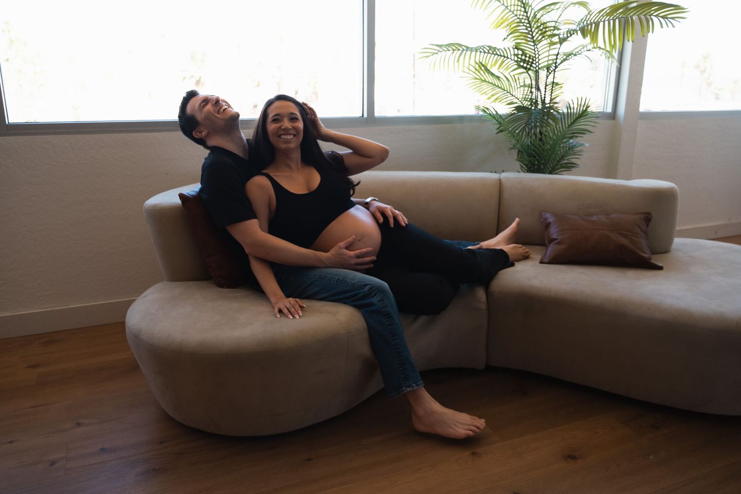 Happy pregnant couple relaxing together on a modern beige sectional sofa in a bright living room.
