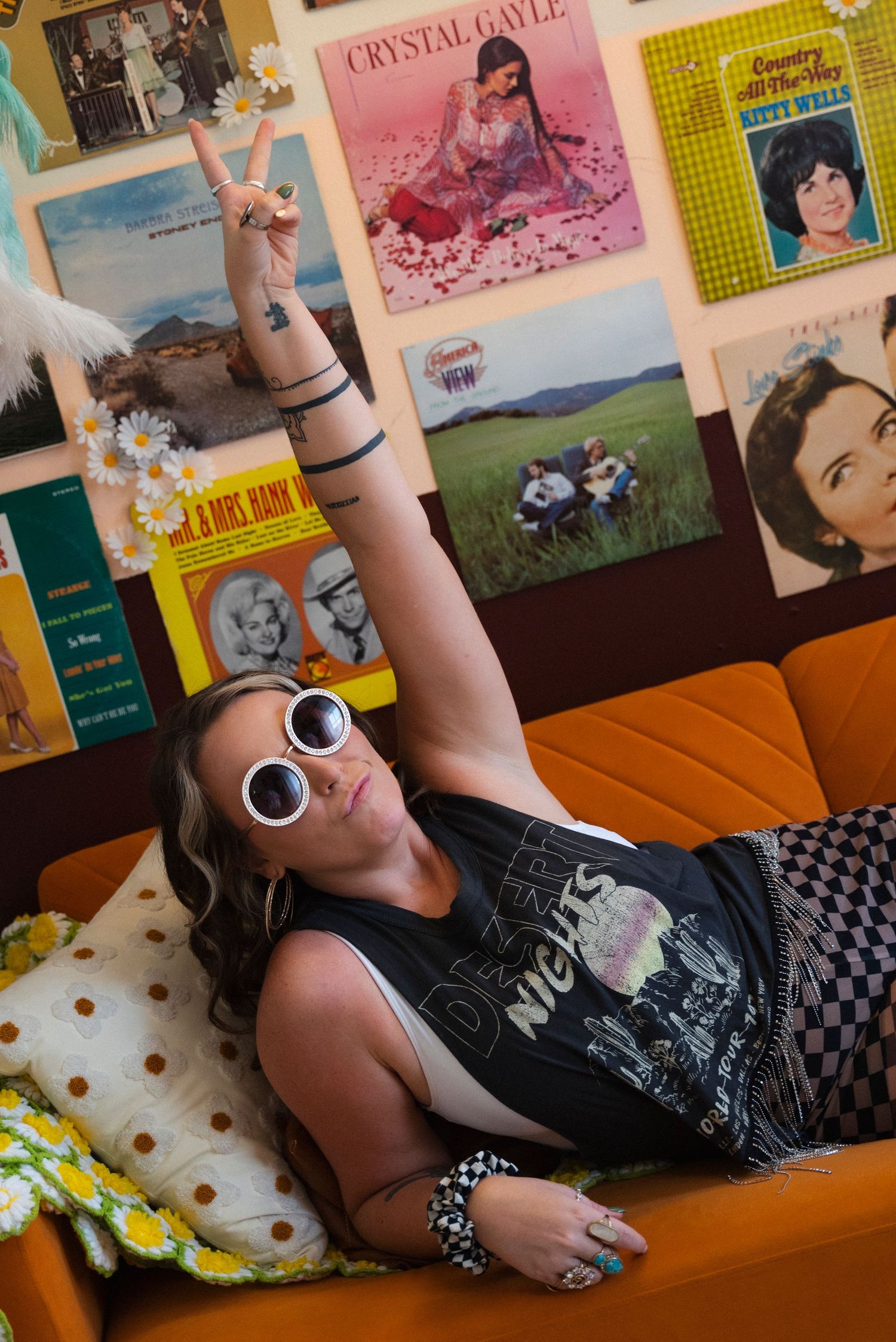Someone in round sunglasses reclines on an orange couch beneath vintage album covers and posters on the wall.