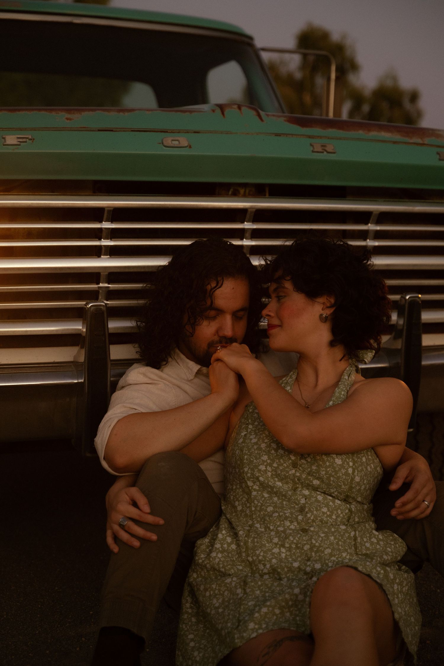 Romantic couple sitting together leaning against a vintage green Ford truck at golden hour.