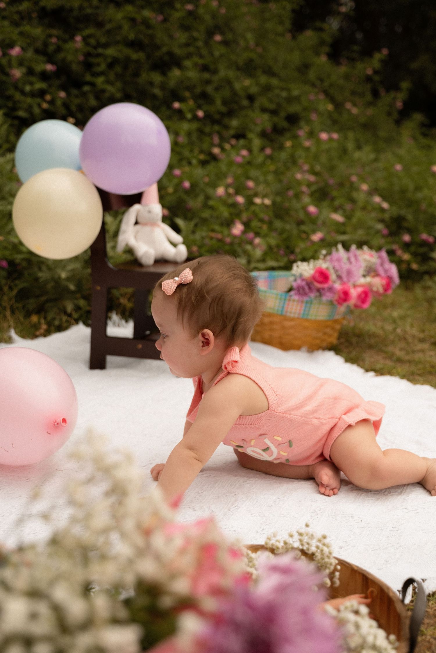 Baby girl in pink romper crawls on white blanket outdoors surrounded by pastel balloons and flowers.