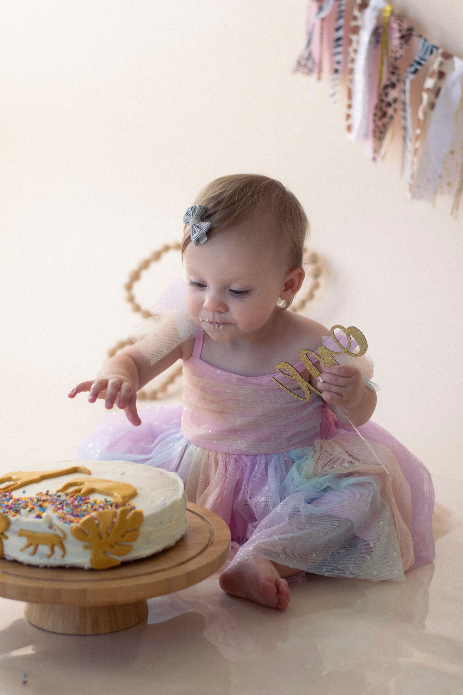 A small child in a pastel tulle dress sits beside a wooden cake stand with decorated pastries.