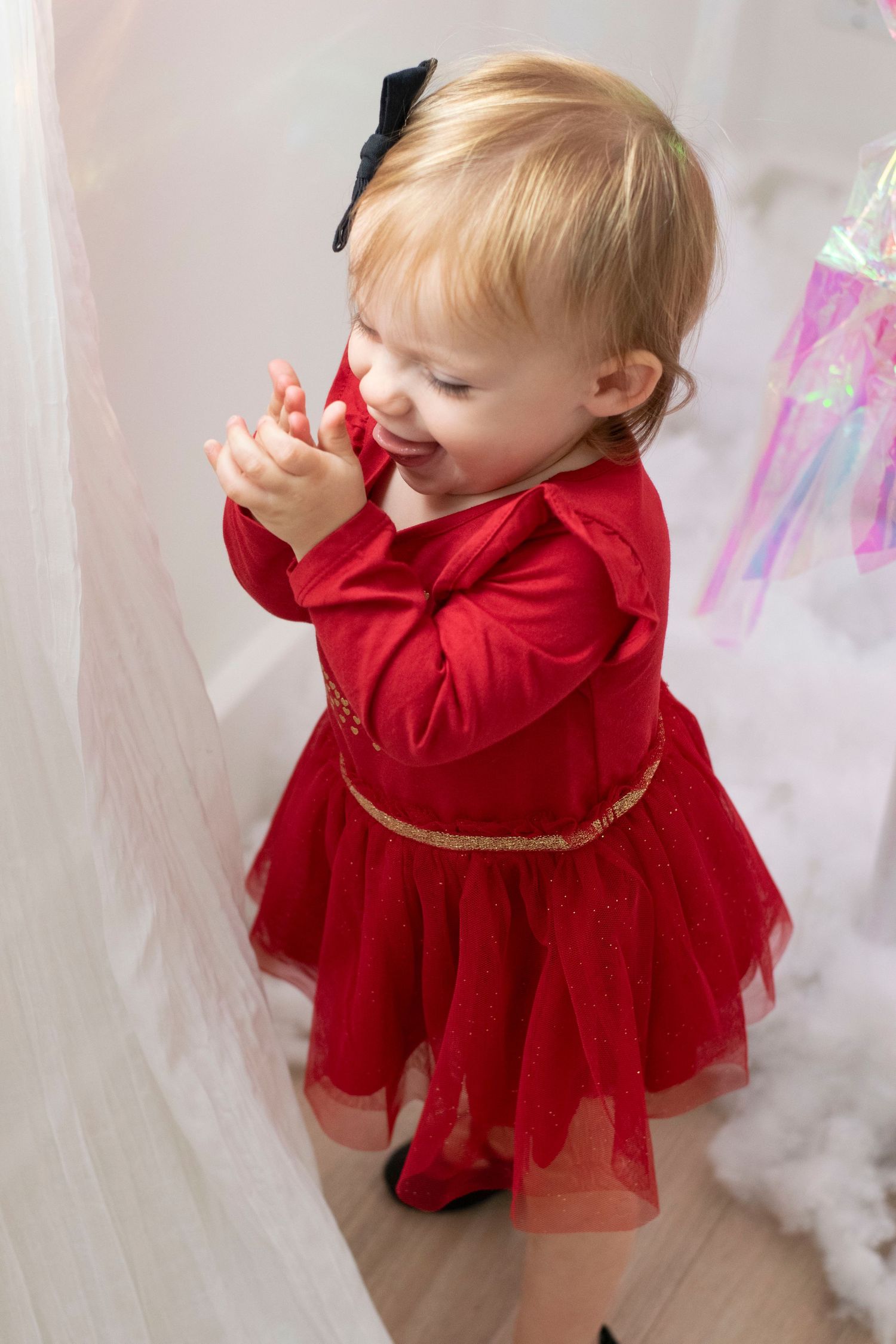 Young toddler wearing a festive red velvet dress with tulle skirt and gold belt stands against white curtains.