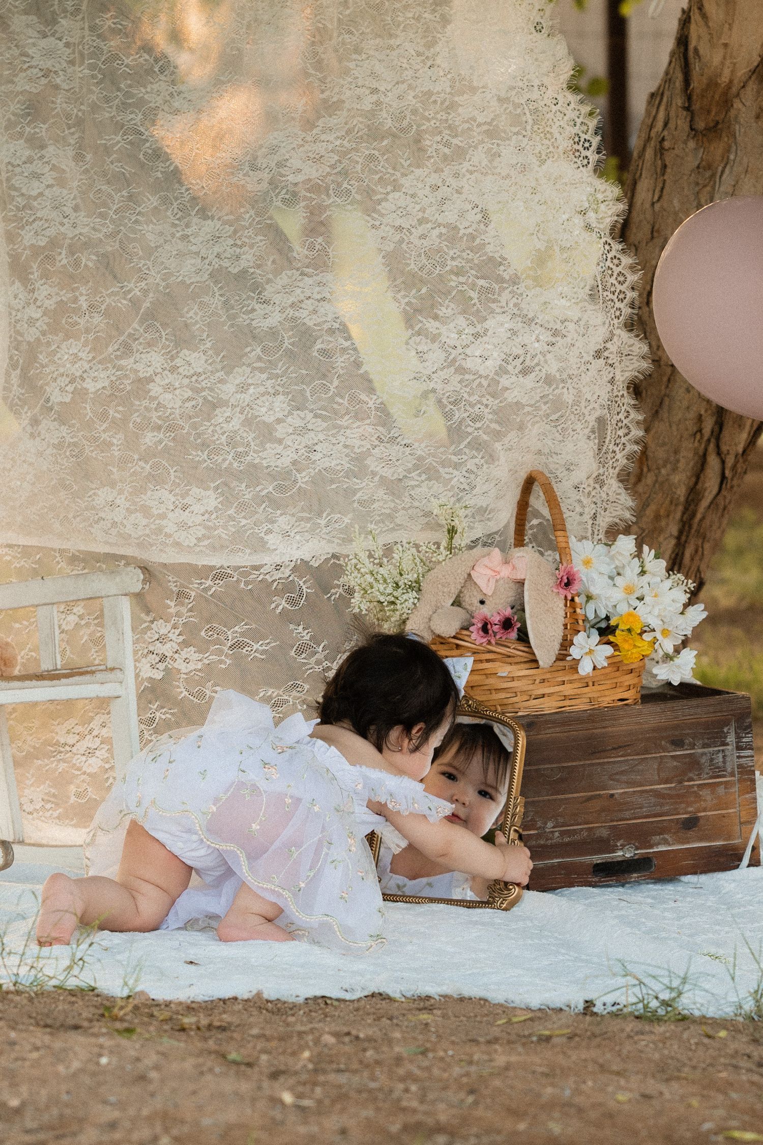 Baby girl in white dress crawling near vintage suitcase with flowers and bunny toy at outdoor photo session.