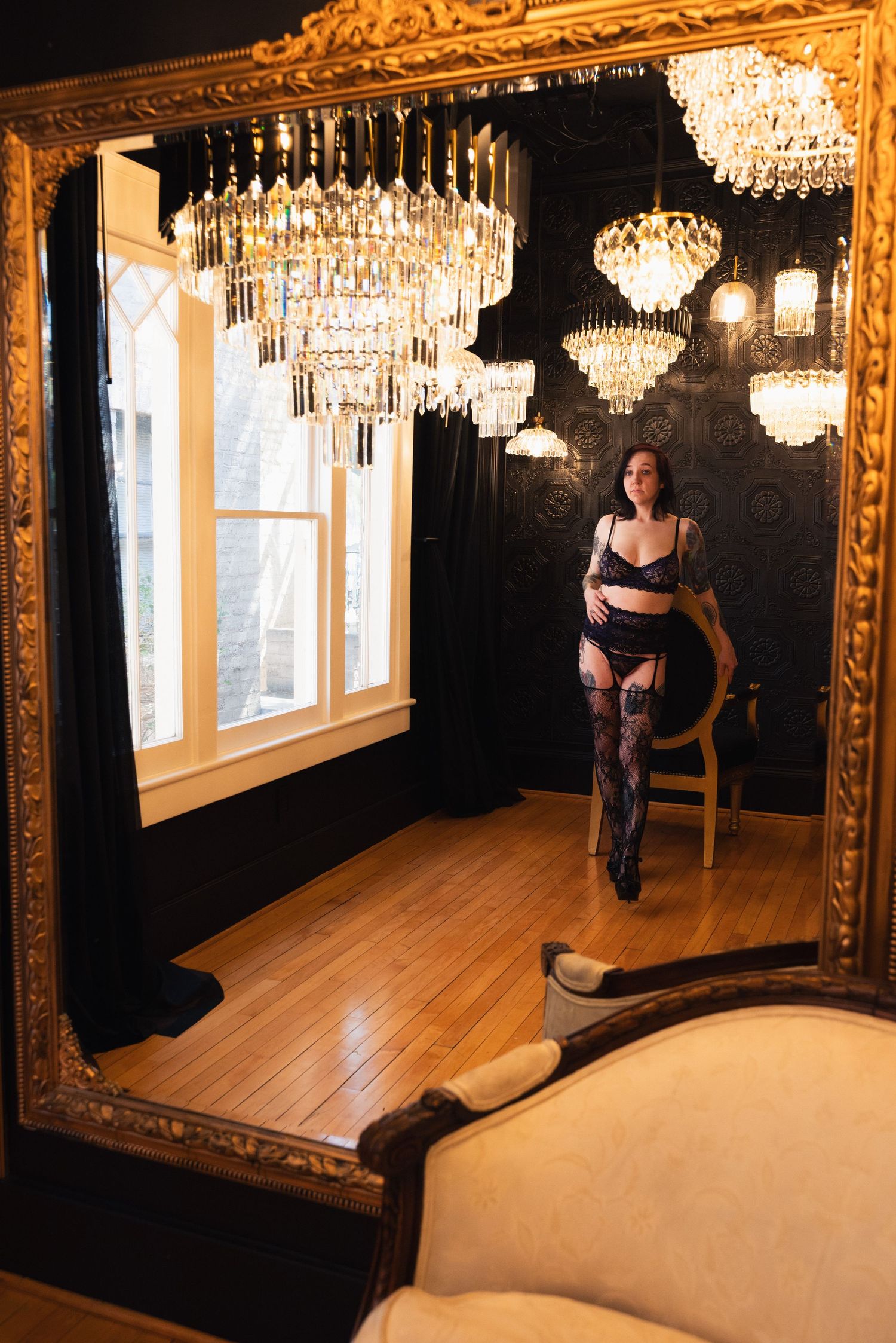 Woman in black lingerie and stockings poses in an ornate gold-framed mirror with crystal chandeliers overhead.