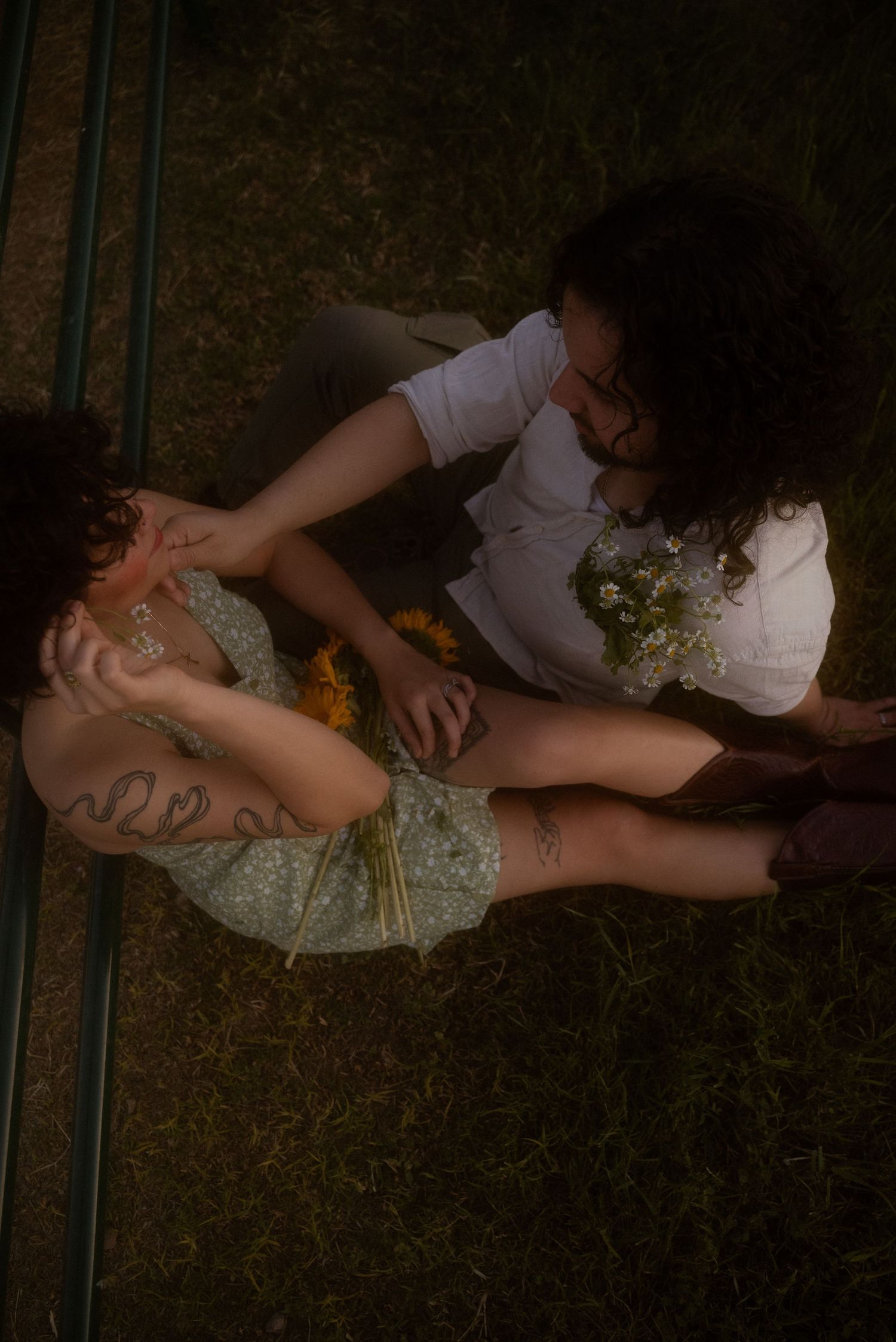 Aerial view of two people sitting on grass, one in a floral dress with tattoos holding wildflowers.