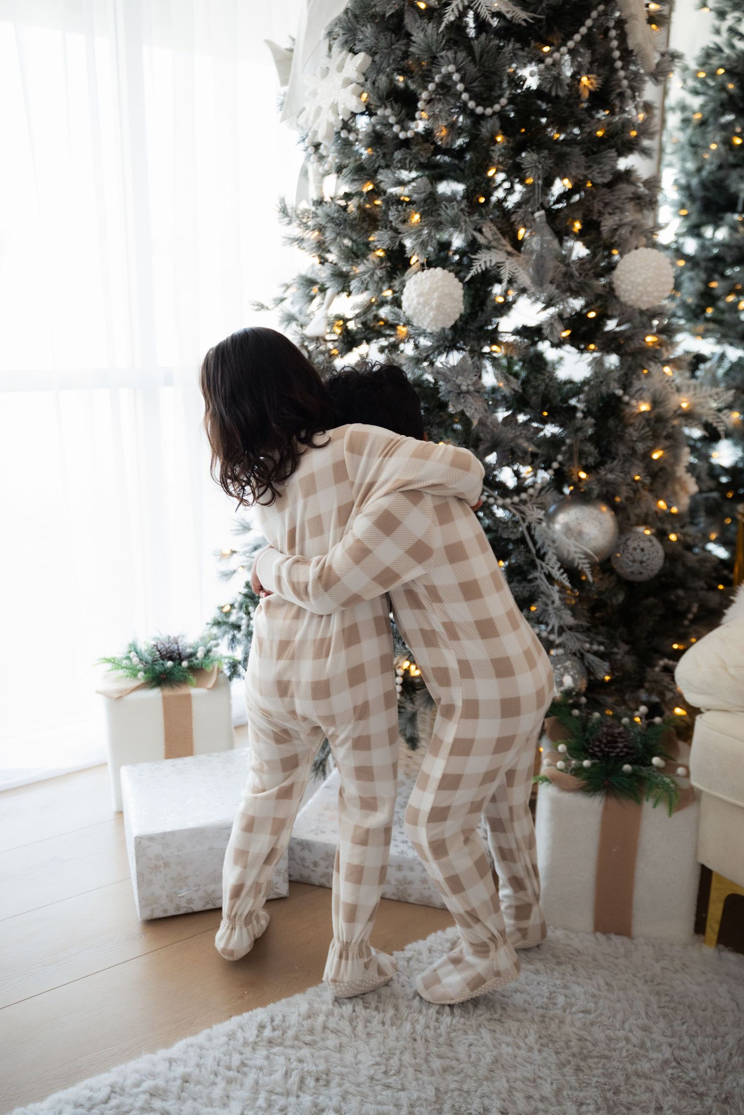 Two children in matching plaid footed pajamas hugging near a decorated Christmas tree with gifts.