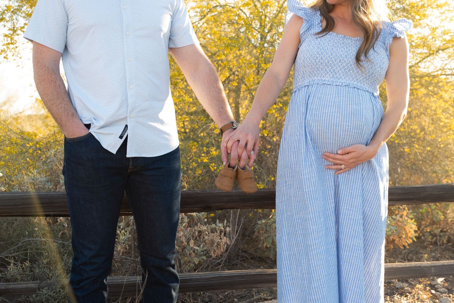 A couple holds hands at sunset, with a pregnant figure wearing a flowing light blue maxi dress against autumn foliage.