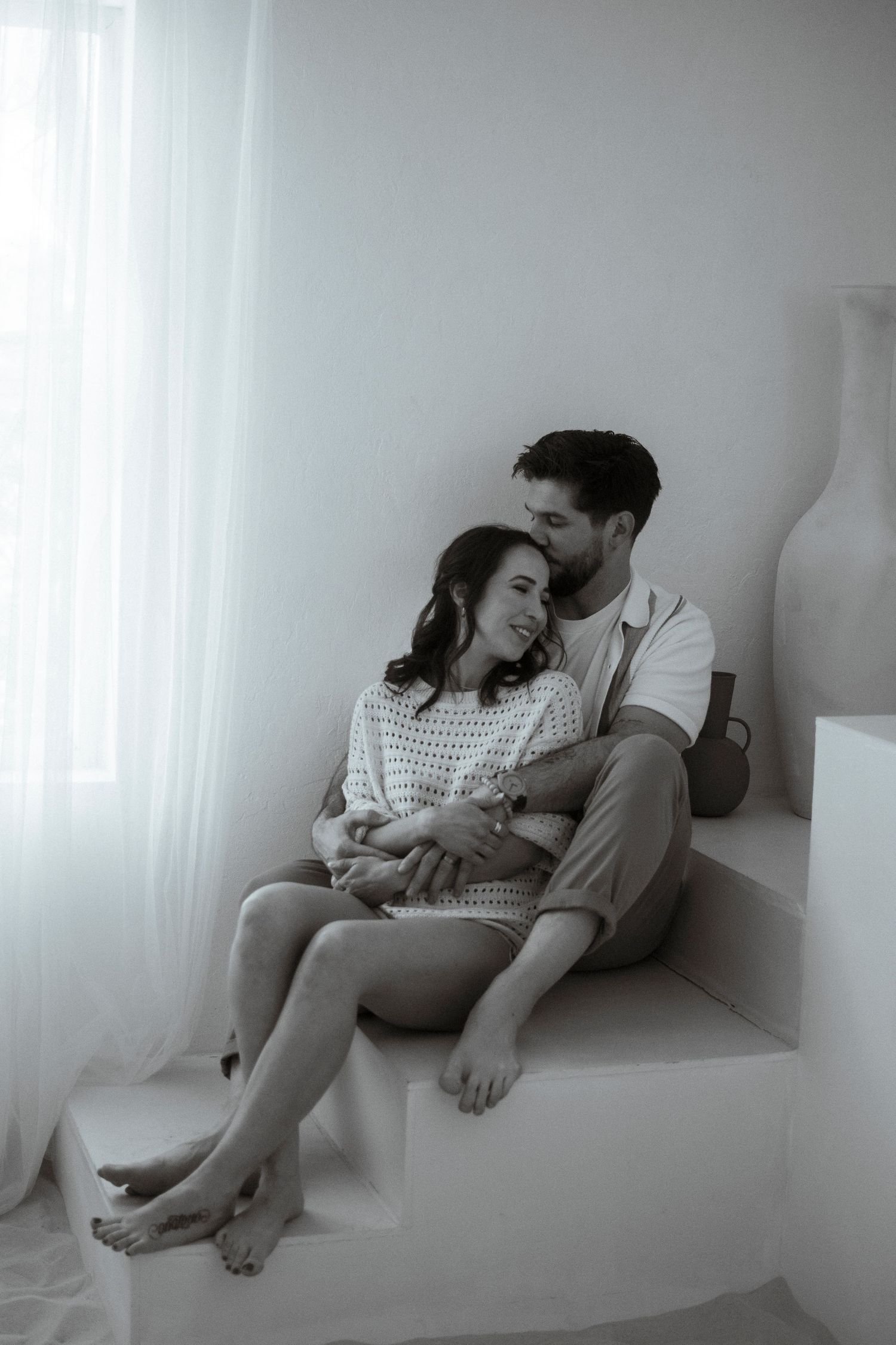 Romantic couple lounging together in white minimalist setting with natural lighting.
