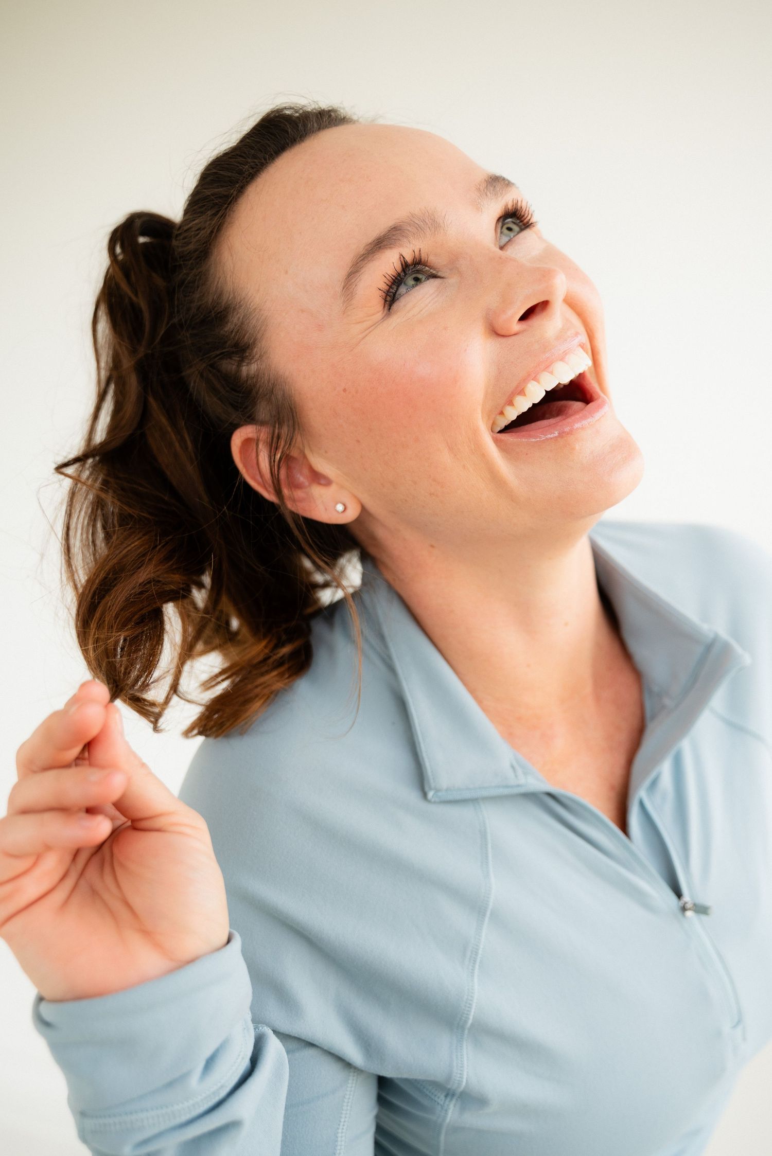 A person in a light blue shirt laughs joyfully while looking upward with their hair pulled back.