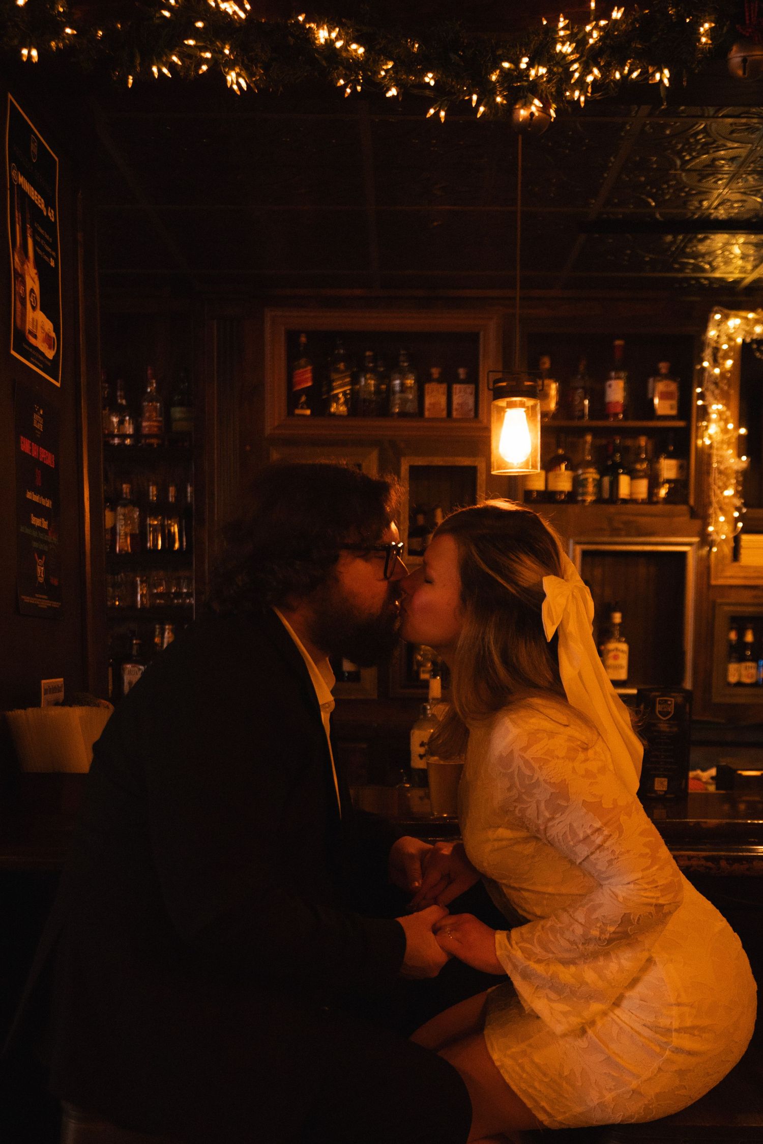 Couple sharing a romantic kiss at a dimly lit bar with warm string lights and liquor bottles in the background.