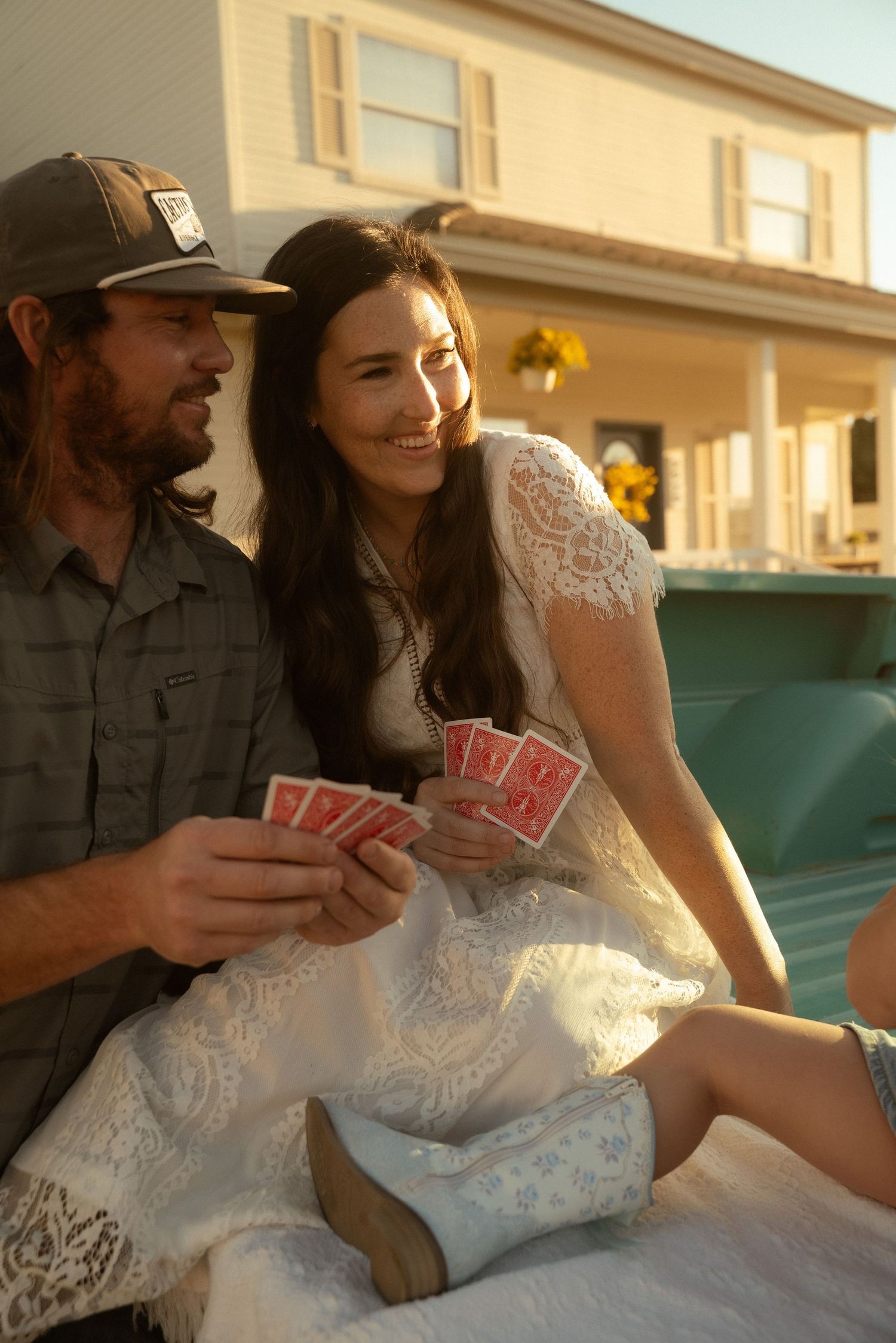 A smiling couple in rustic attire plays cards outdoors near a farmhouse at golden hour, bride in lace dress and boots.