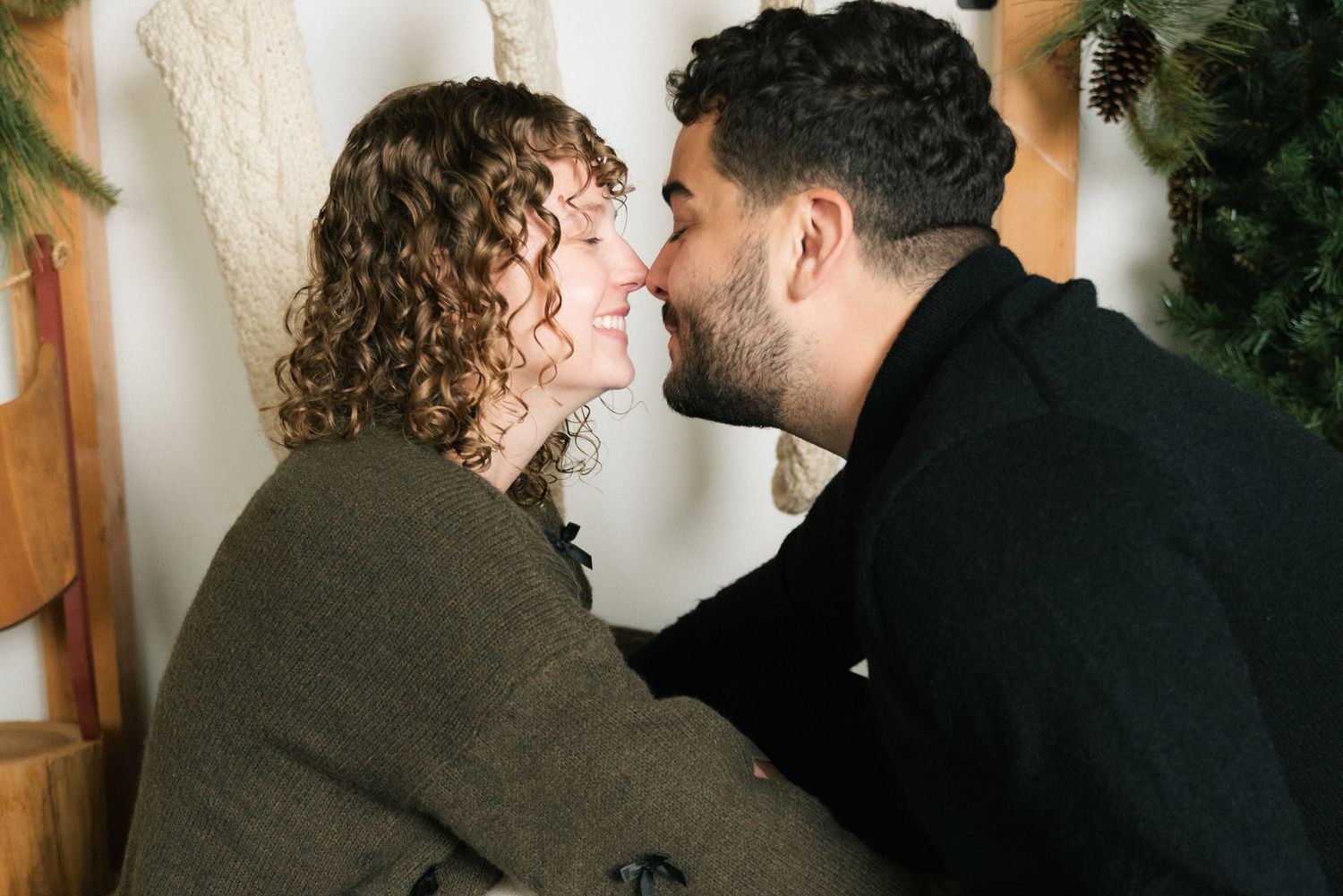 Two people share an intimate moment in front of a white wall with green foliage visible in the background.