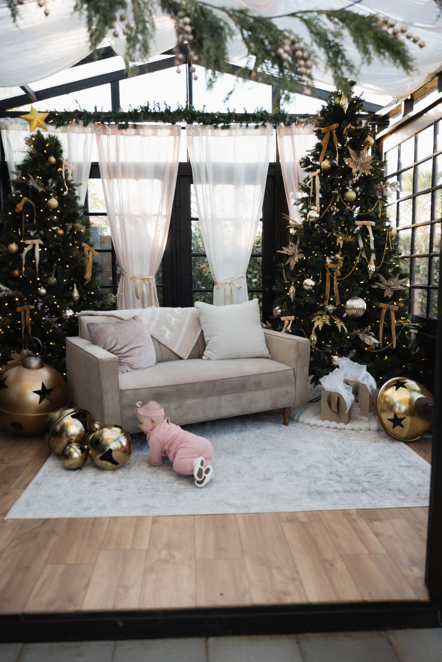 A cozy sunroom decorated with Christmas trees, white cushions, and gold ornaments creates an elegant holiday atmosphere.