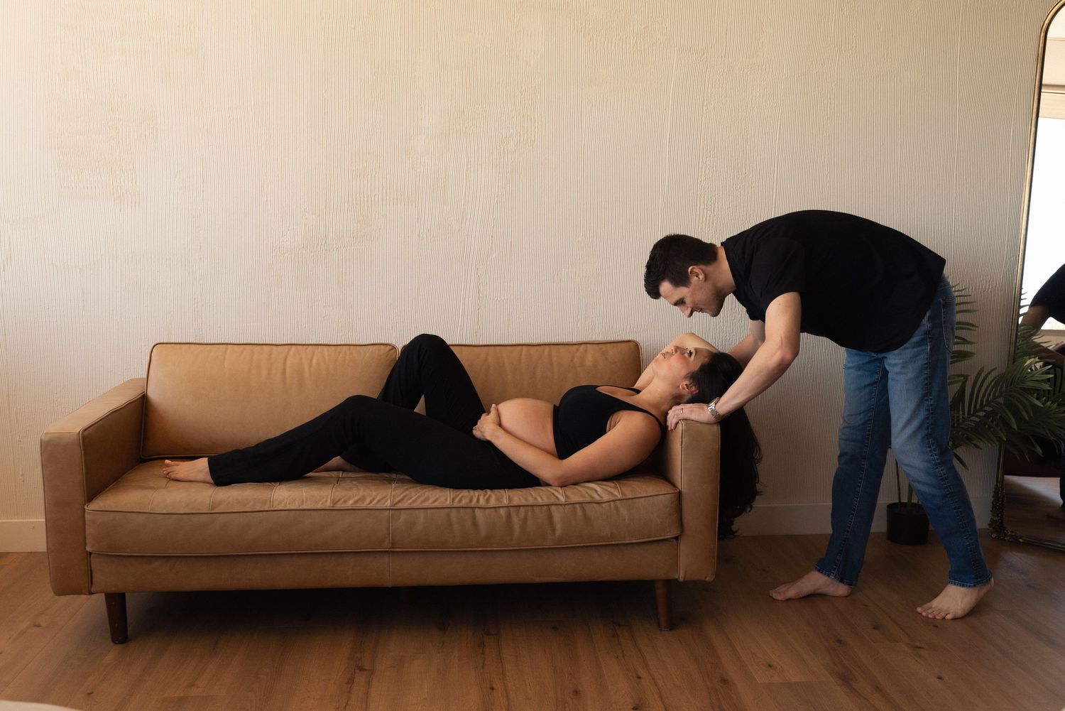 Pregnant woman lying on brown sofa while partner leans over her in a tender moment at home.