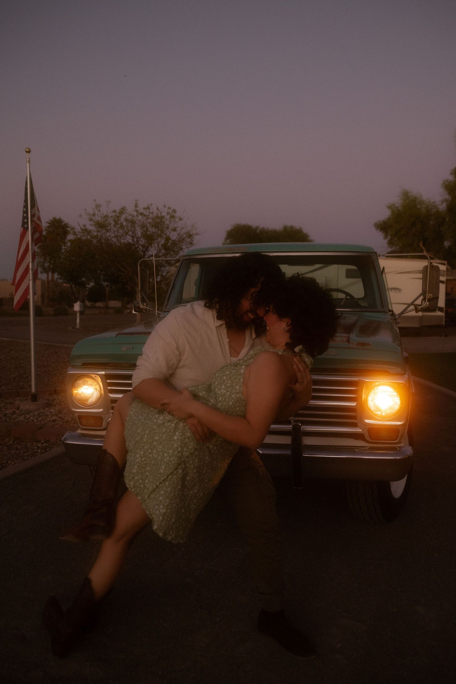 A romantic couple shares a kiss leaning against a vintage truck at dusk with an American flag in the background.