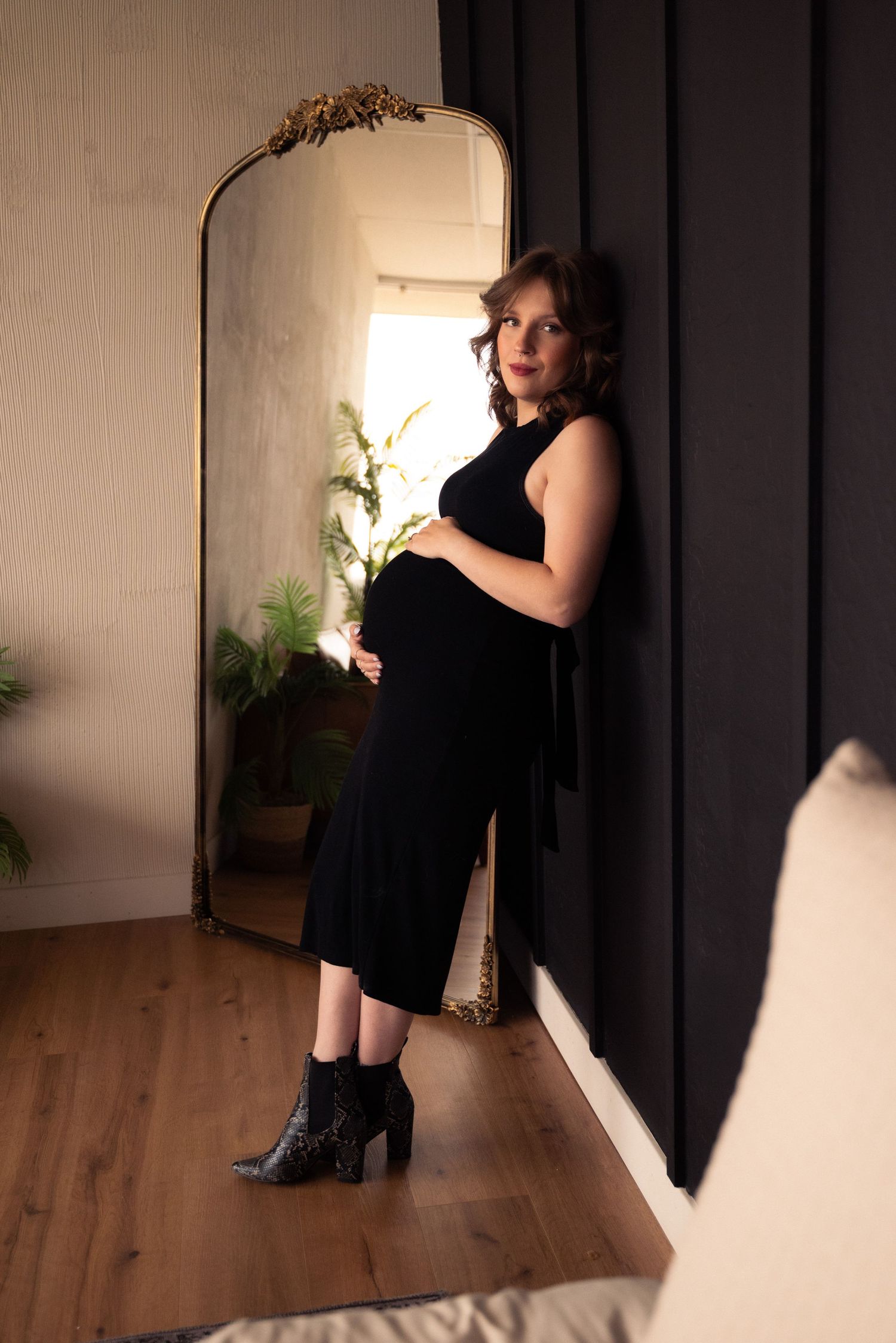 Expectant mother in elegant black dress poses near vintage mirror and houseplants on hardwood floor.