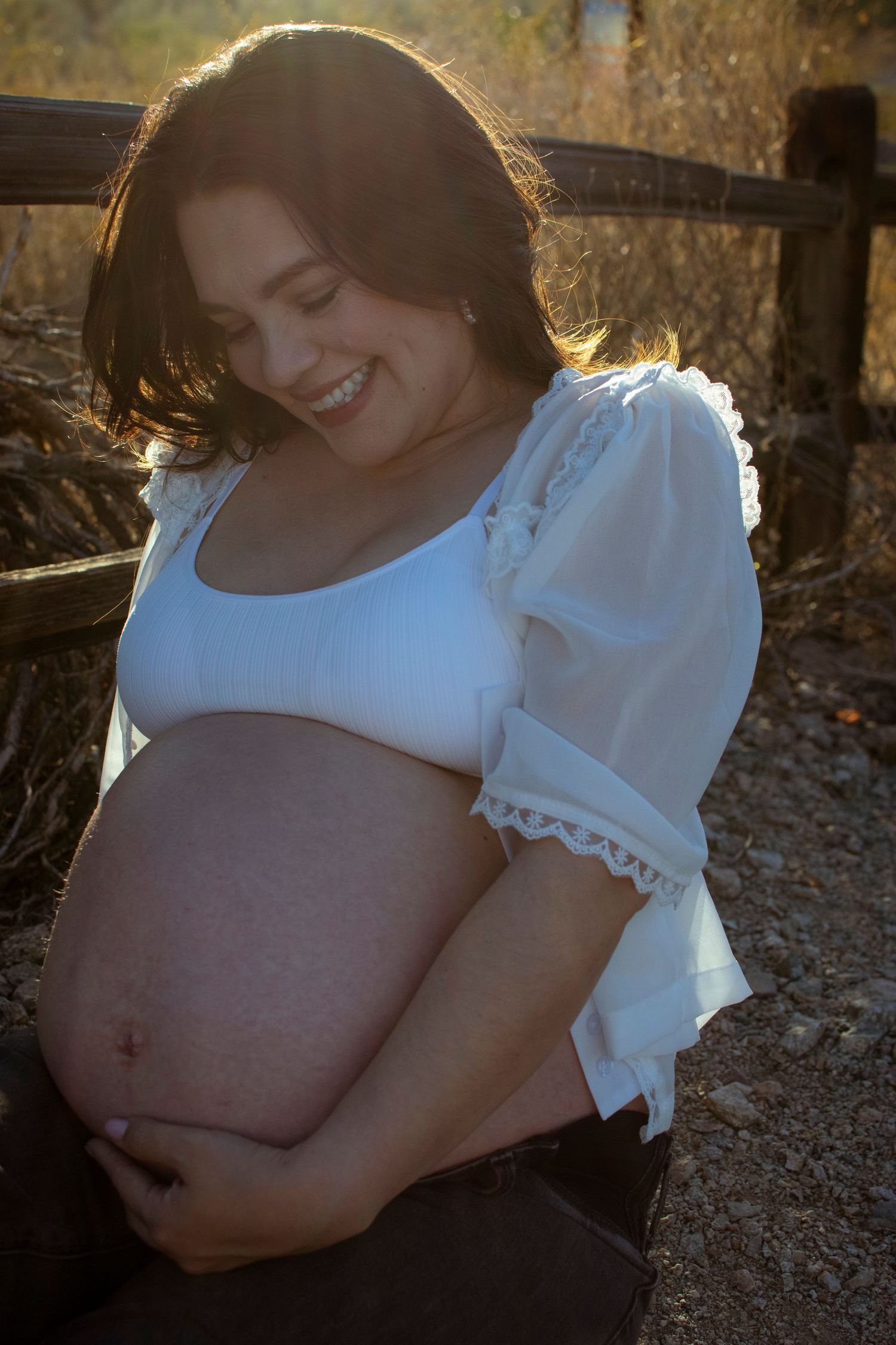 A pregnant woman in a white lace dress smiles while sitting on a wooden bench at sunset.