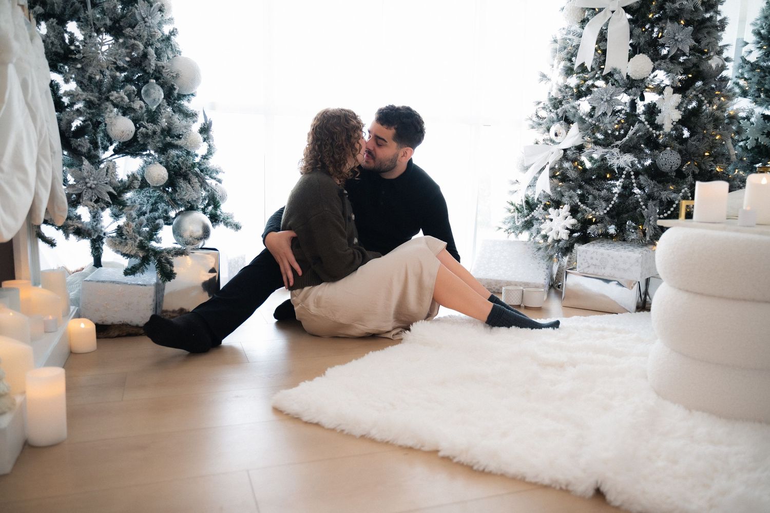 Couple sits together on white fur rug in front of decorated Christmas trees in bright modern room.