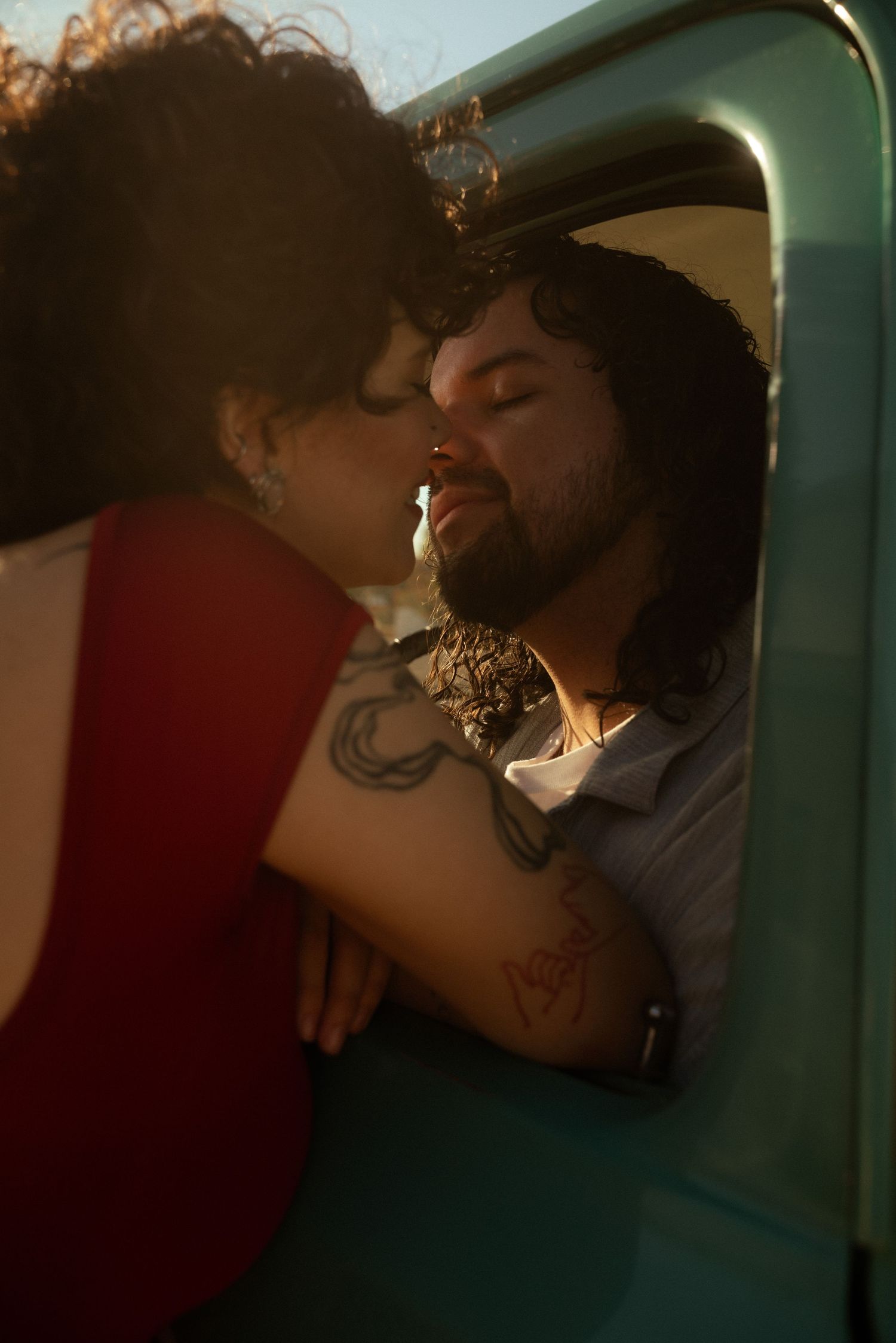 A couple shares a romantic kiss through a vintage truck window in warm golden sunlight, the man showing tattoos on his arm.