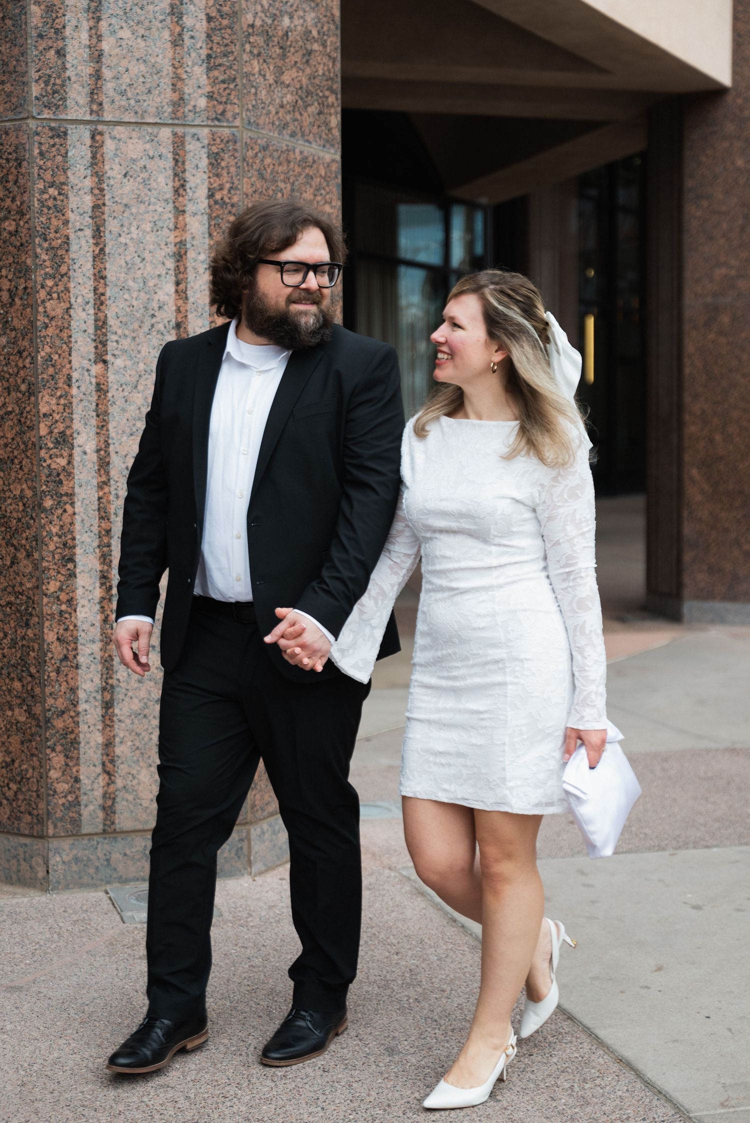 Couple in contrasting black and white formal attire playfully pose together against a stone column backdrop.