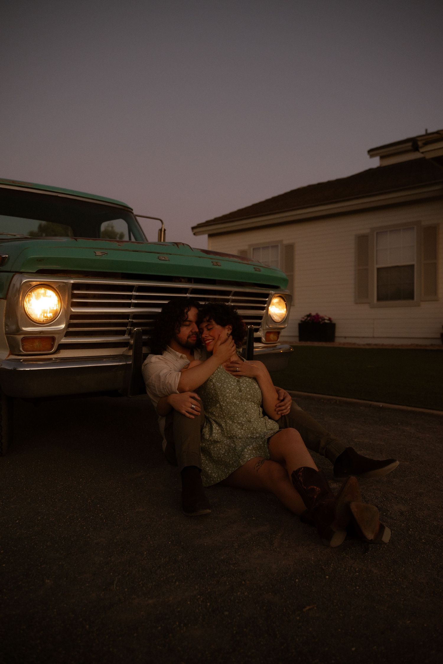Couple sitting together in front of vintage truck headlights at dusk, romantic evening outdoor scene.
