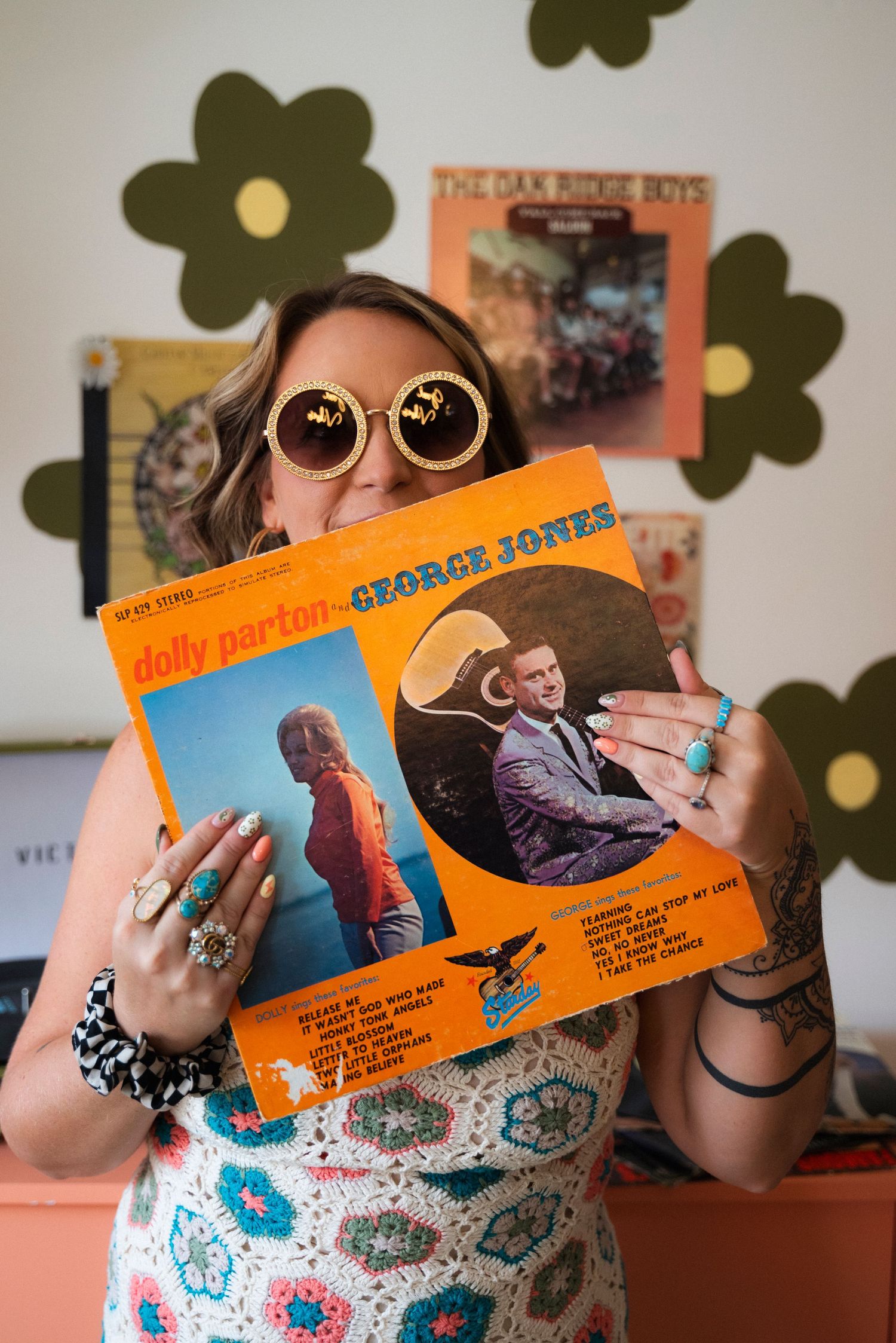 Person in floral dress holding vinyl records displaying orange album covers against a patterned background.