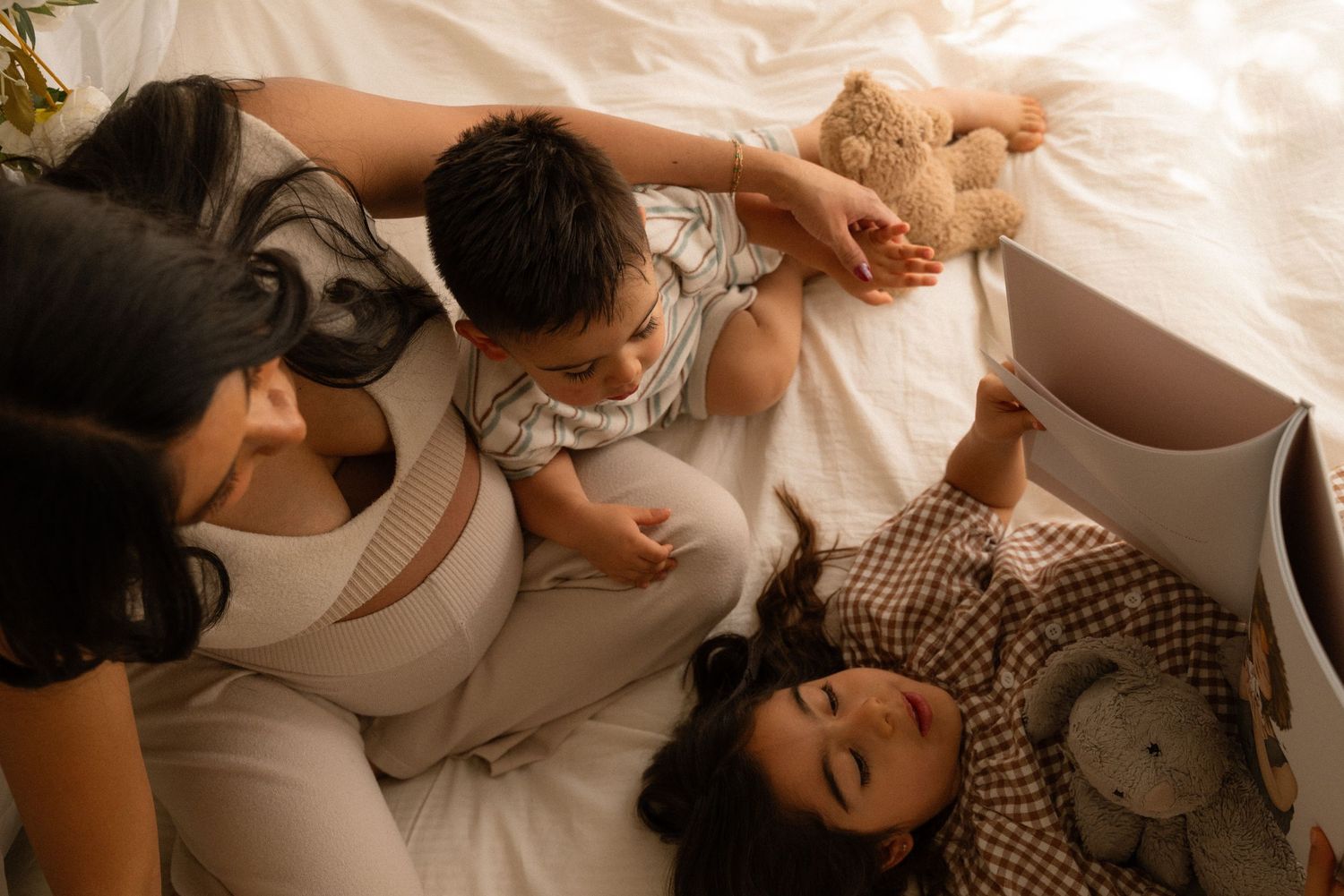A mother and two young children relax together on a white bed with stuffed animals in a cozy, warmly lit room.