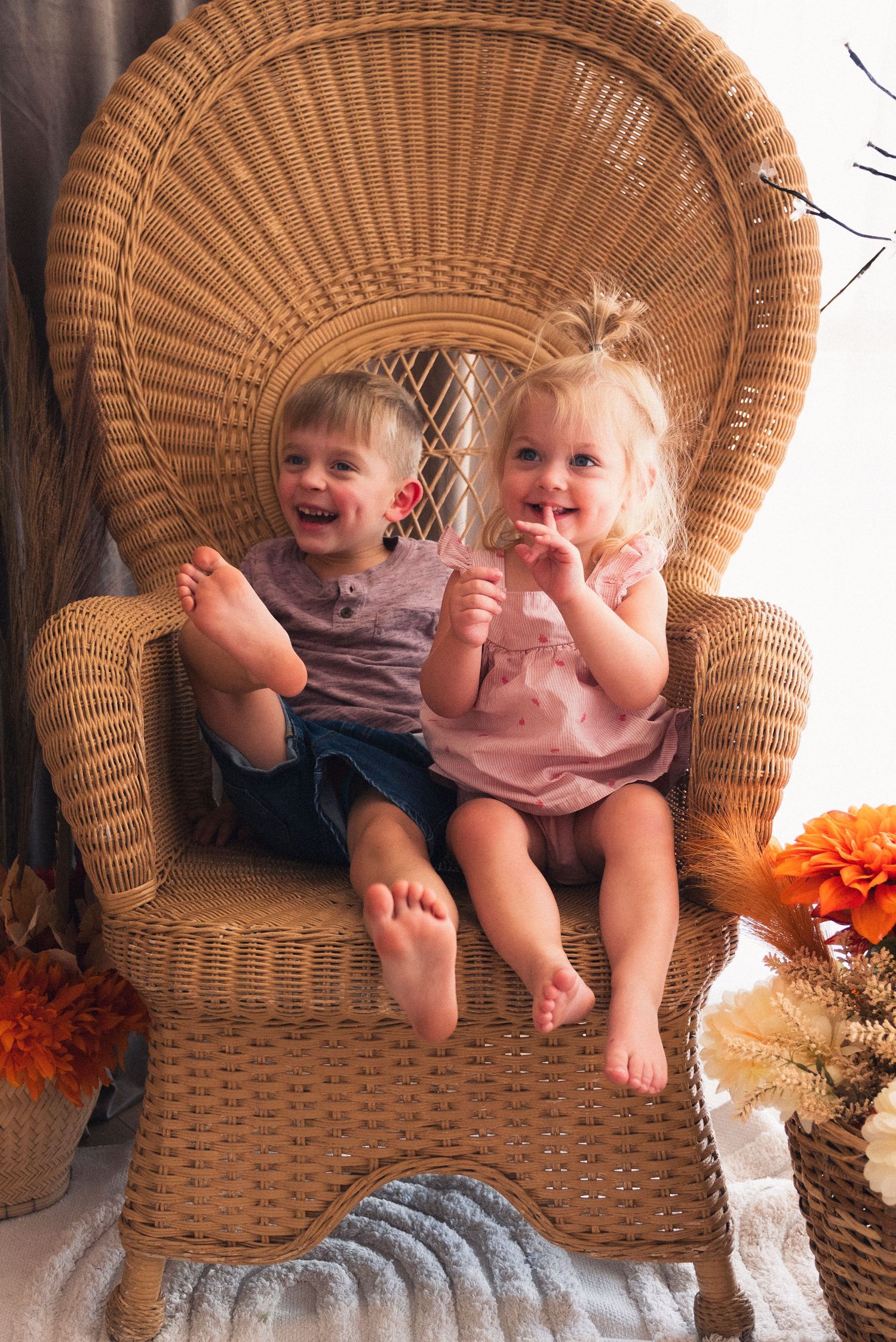 Two children sit together in a woven wicker chair surrounded by autumn decorations and fall foliage.