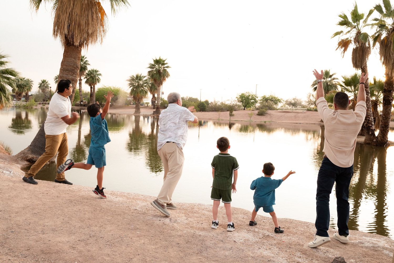 Multi-generational family throwing rocks into a calm lake surrounded by palm trees outdoors.