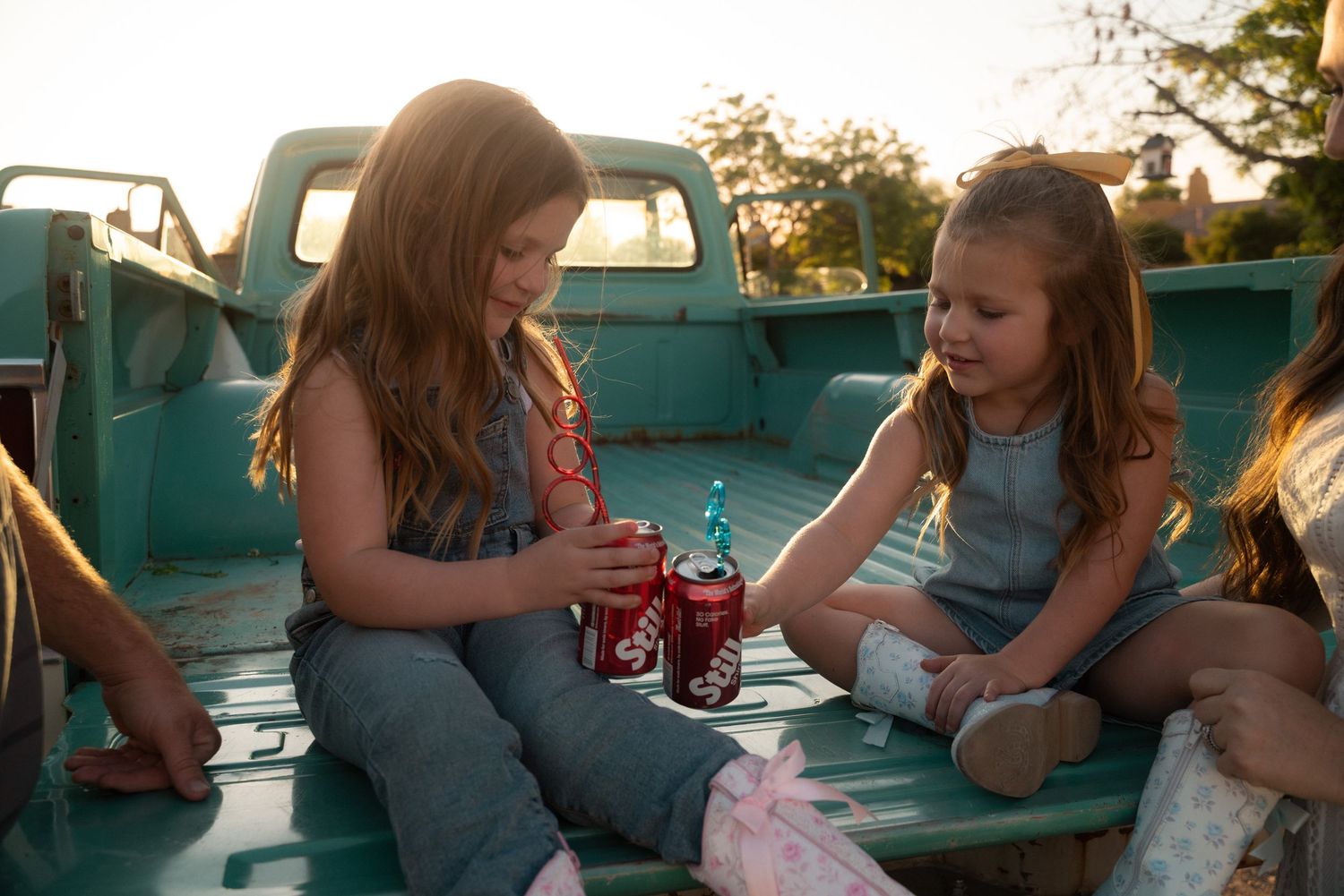 Two young girls sitting in a vintage teal truck bed sharing Coca-Cola drinks in warm sunset light.