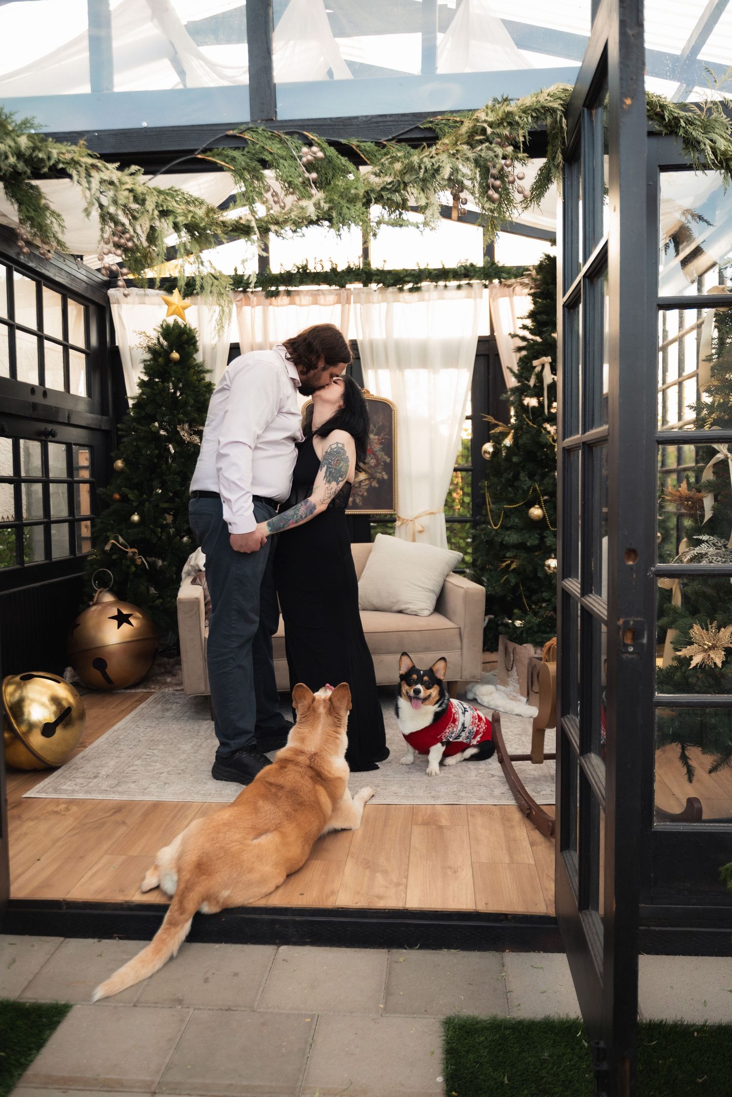 Couple kissing in a festive glass greenhouse decorated with Christmas trees, garland, and two dogs nearby.