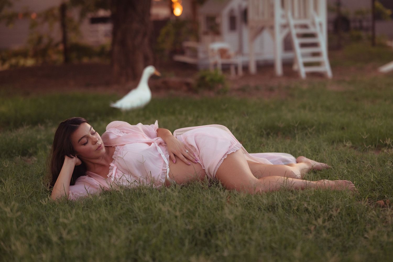 Pregnant woman in pink dress lying on green grass at dusk with a white duck in the background.