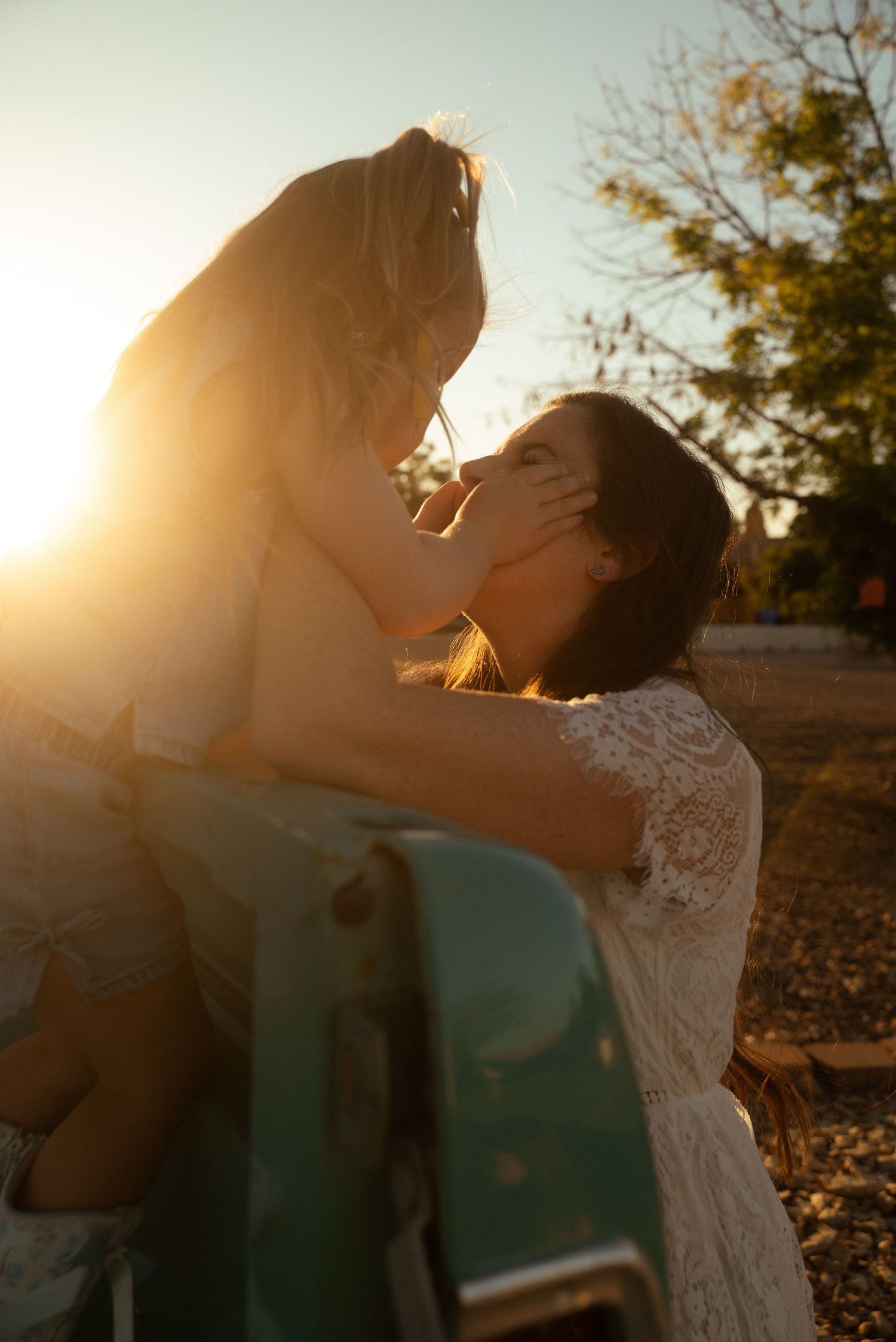 Woman in lace dress kissing her dog at golden hour sunset outdoors near vintage teal car.