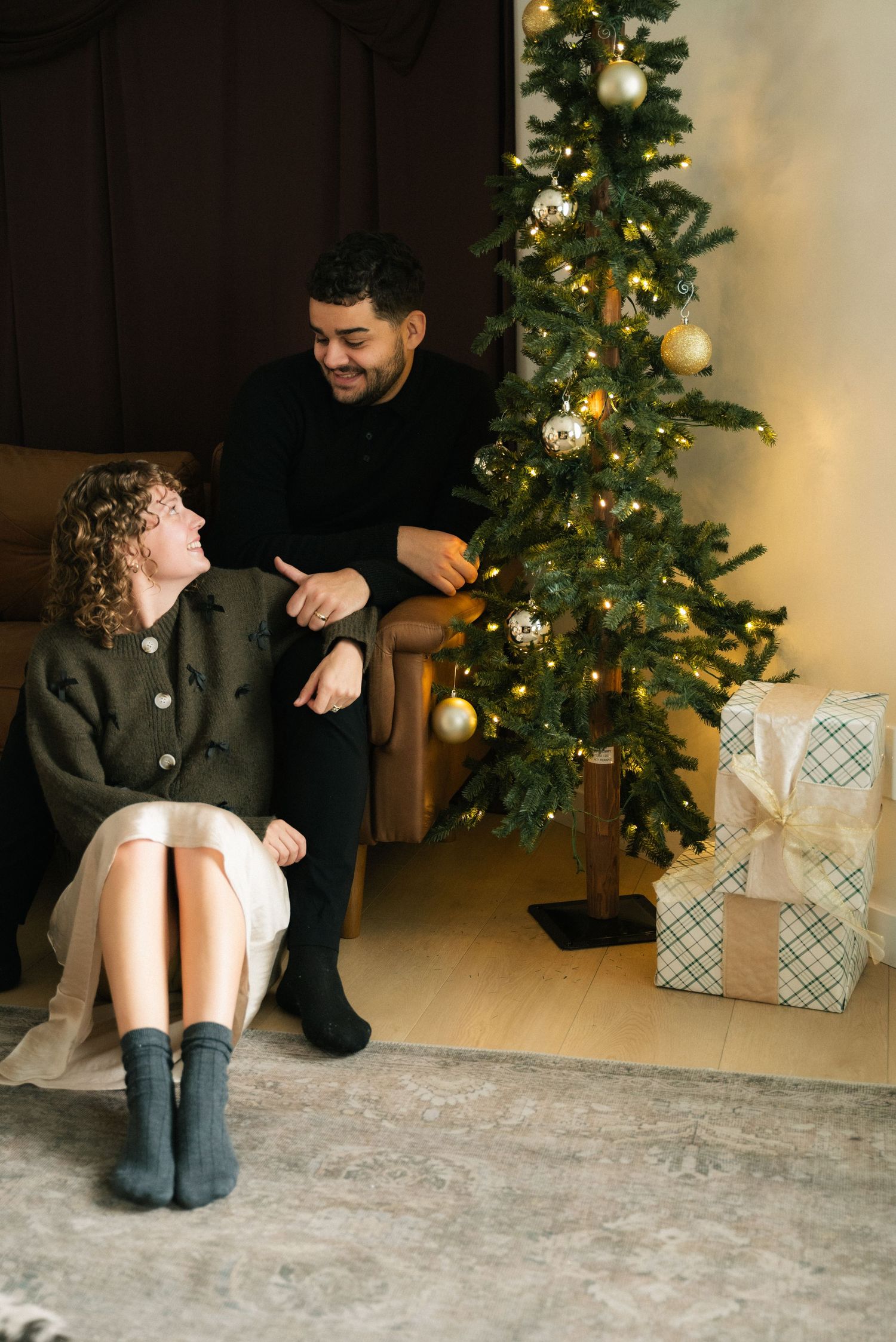 A cozy Christmas scene with two people sharing a joyful moment by a decorated tree with wrapped presents nearby.