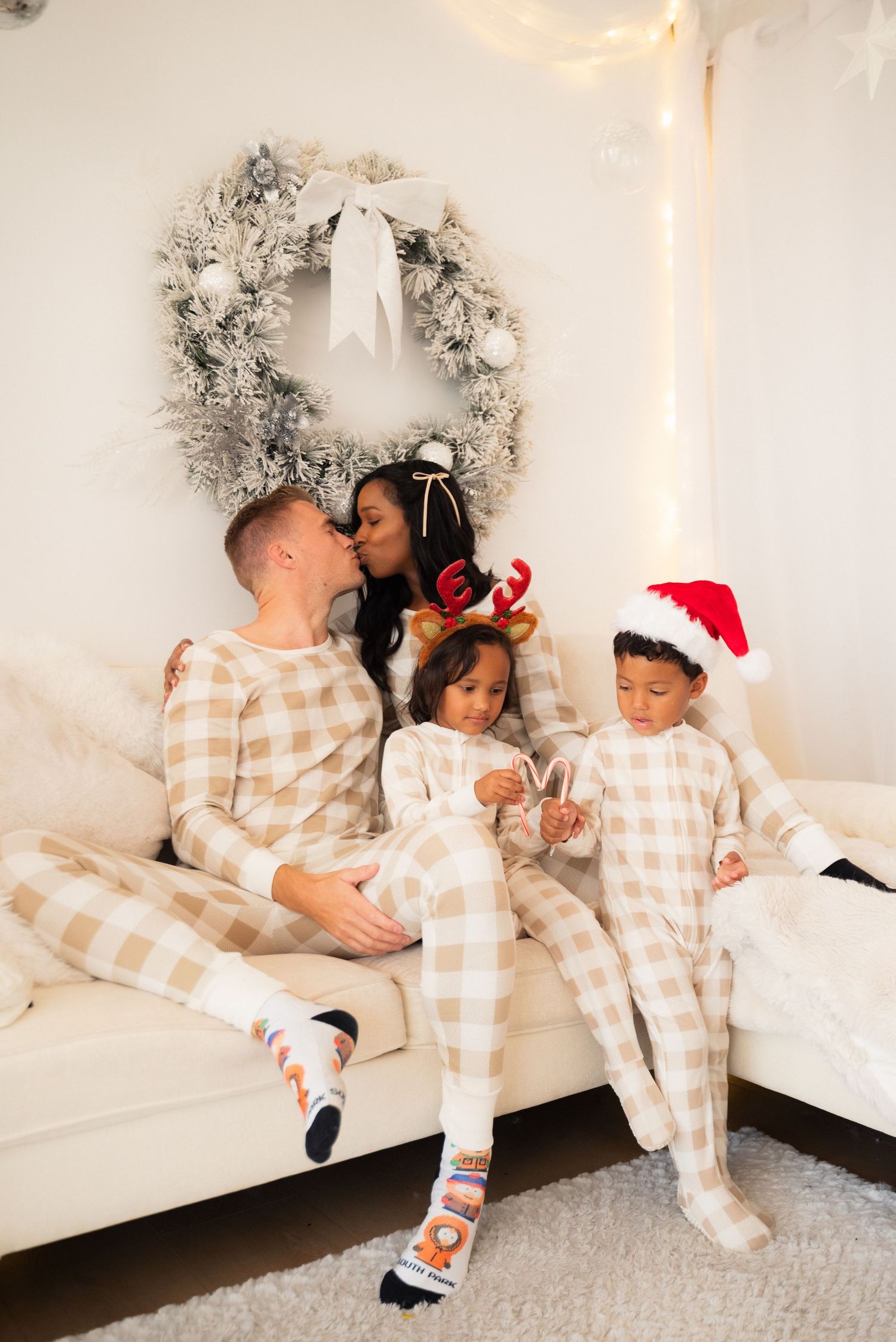 Family in matching plaid Christmas pajamas on a white couch with a holiday wreath, kids in festive headwear.