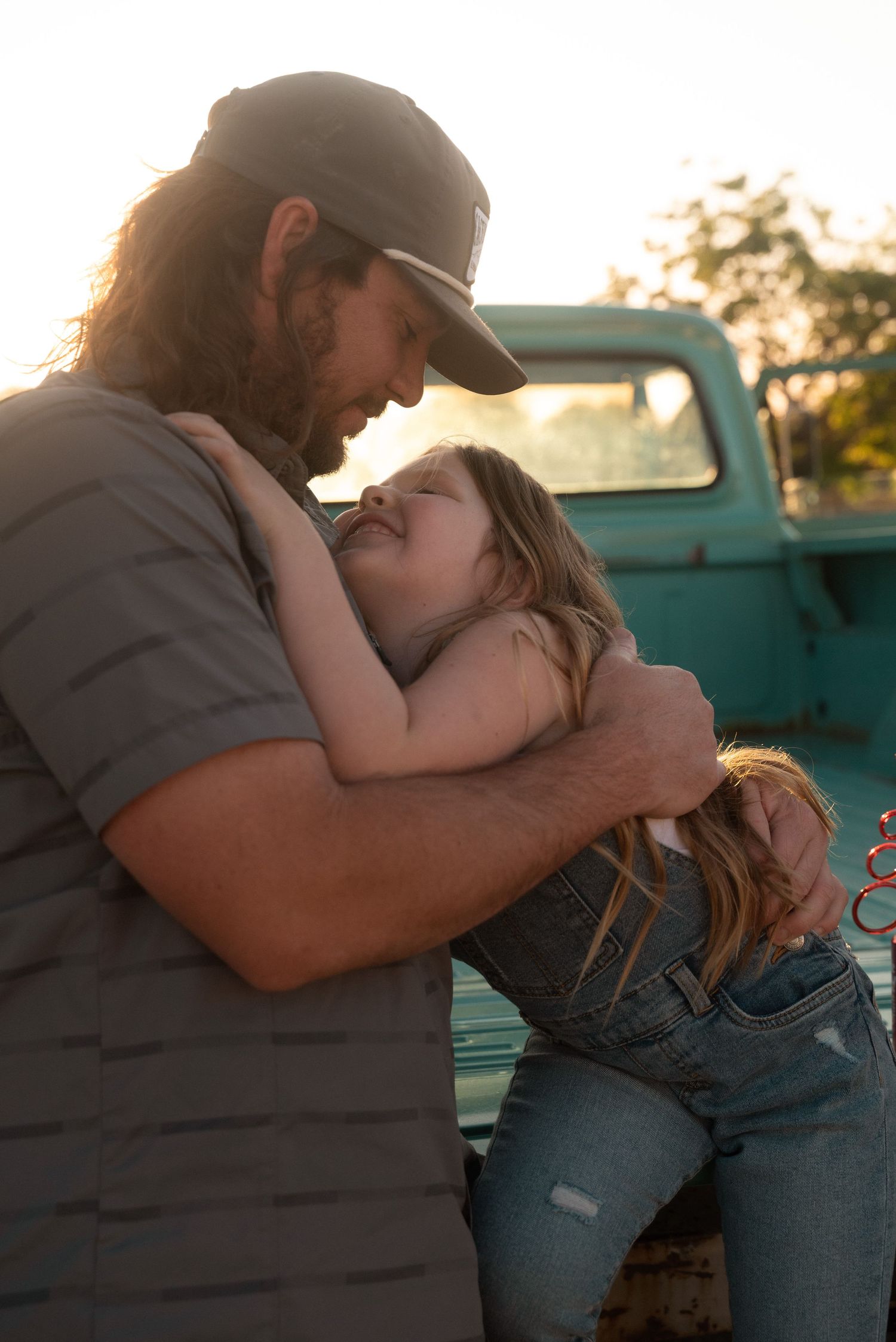 Bearded man in baseball cap tenderly embracing young girl near vintage teal truck at golden hour.