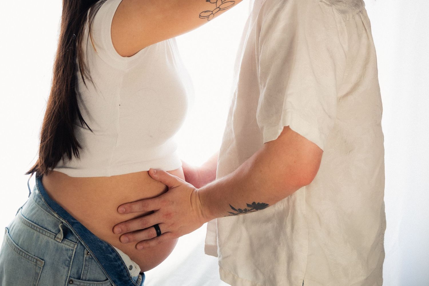 A pregnant woman in white dress cradles her belly while standing in soft natural light.