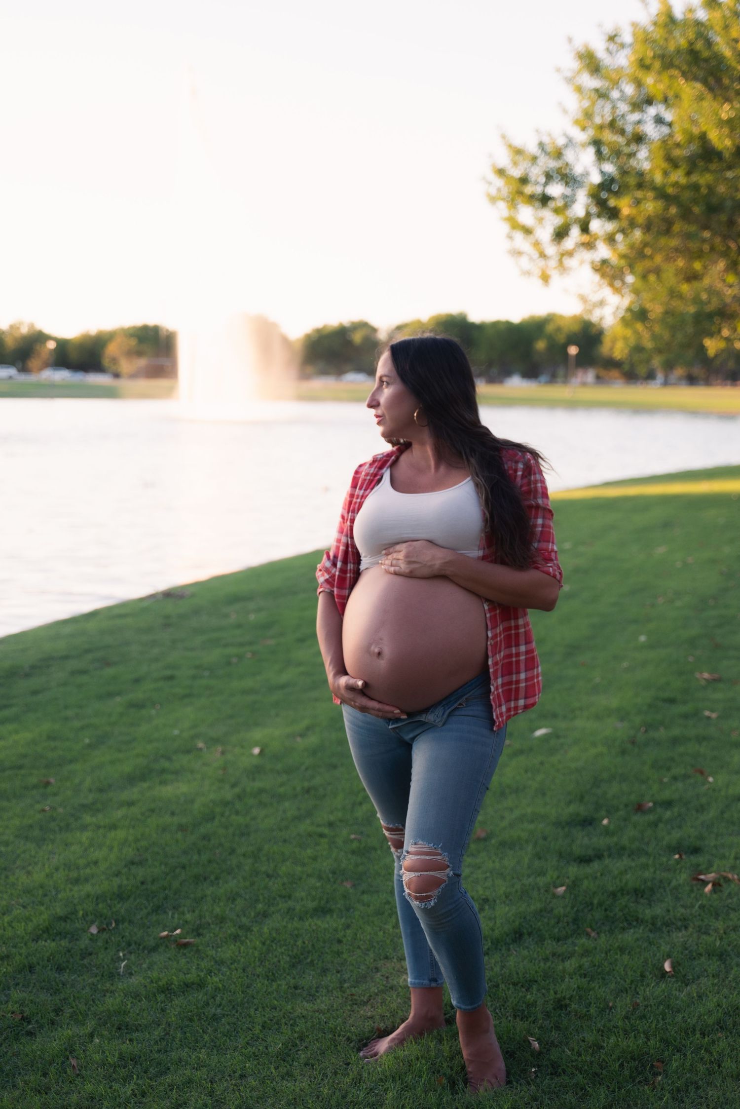 A pregnant person in a plaid shirt and jeans poses outdoors on a grassy lawn at sunset.