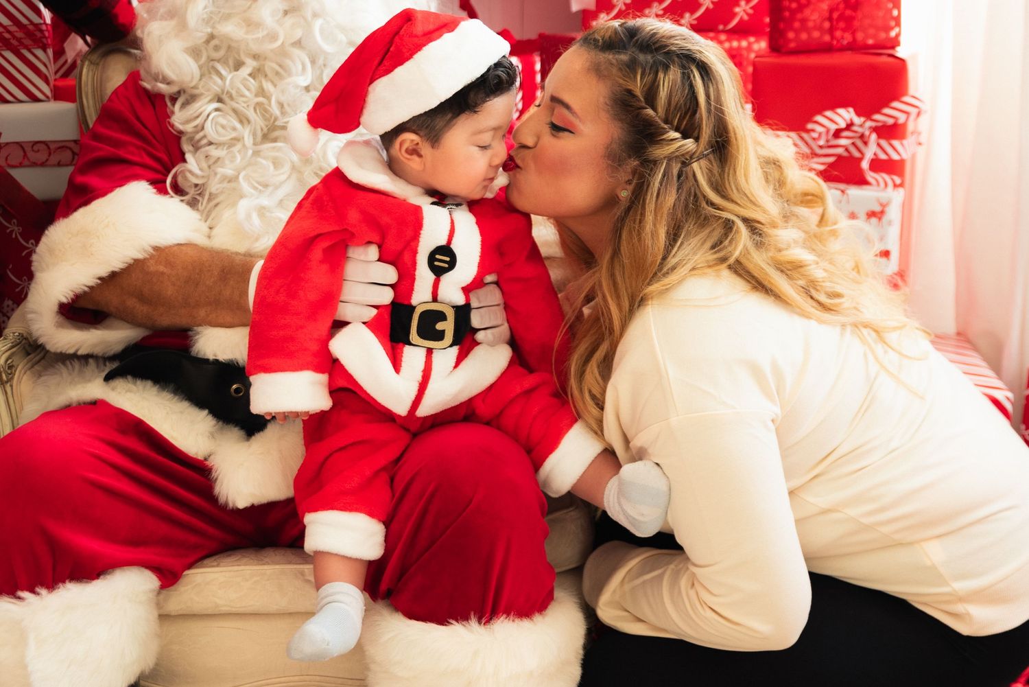 A woman in white kisses a baby dressed in a Santa suit while sitting on Santa's lap at Christmas.