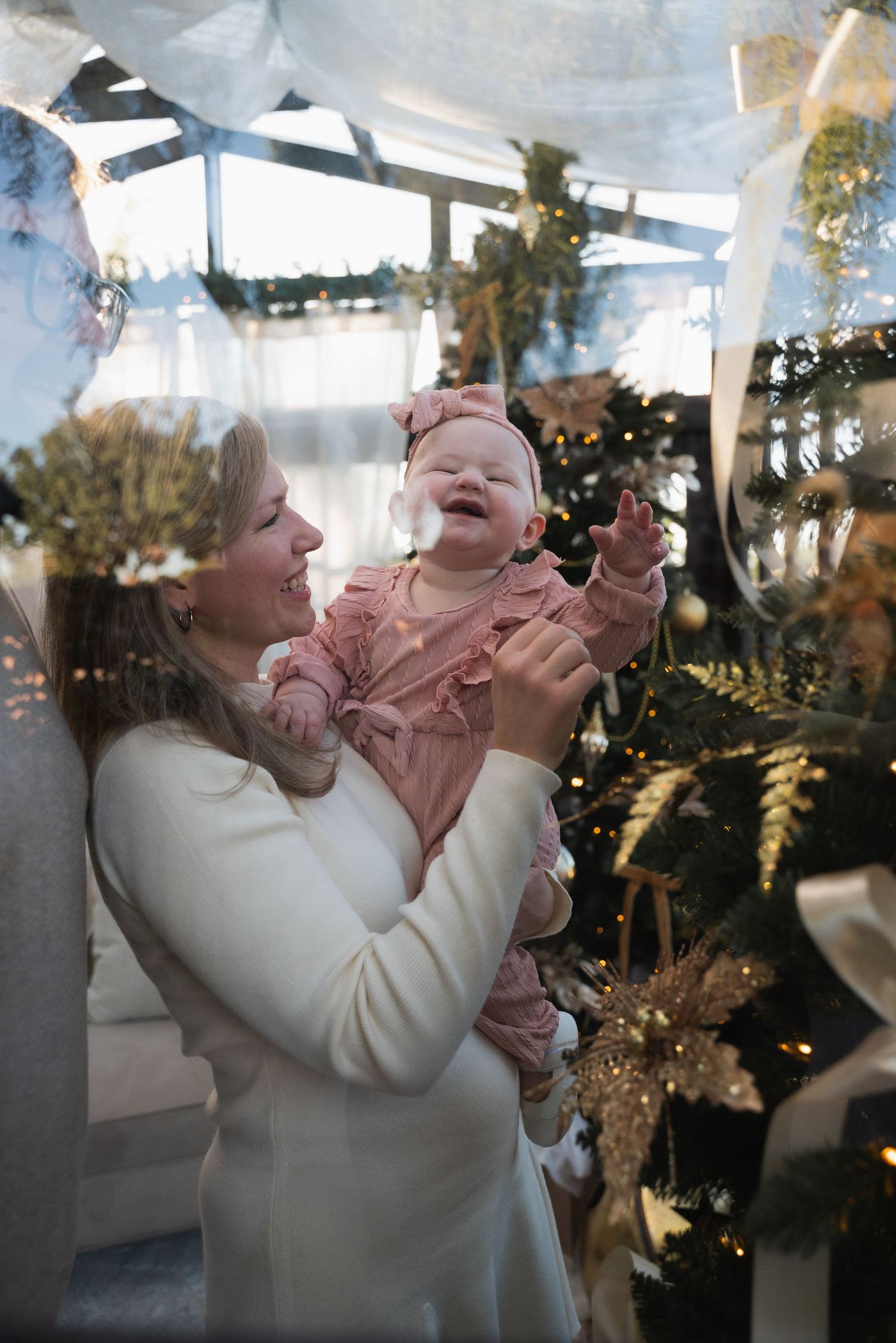 A joyful woman in white holds a laughing baby in pink near a decorated Christmas tree indoors.